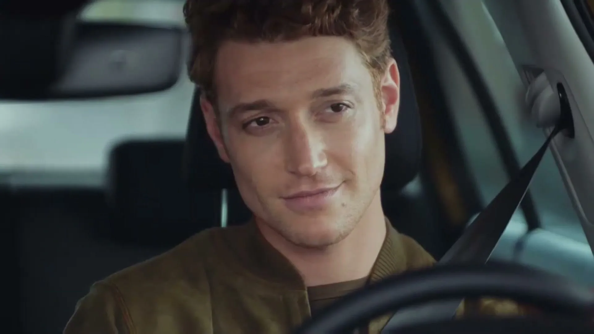 A young man with curly red hair, wearing a green jacket, sits in the driver's seat of a car, looking forward with a slight smile.
