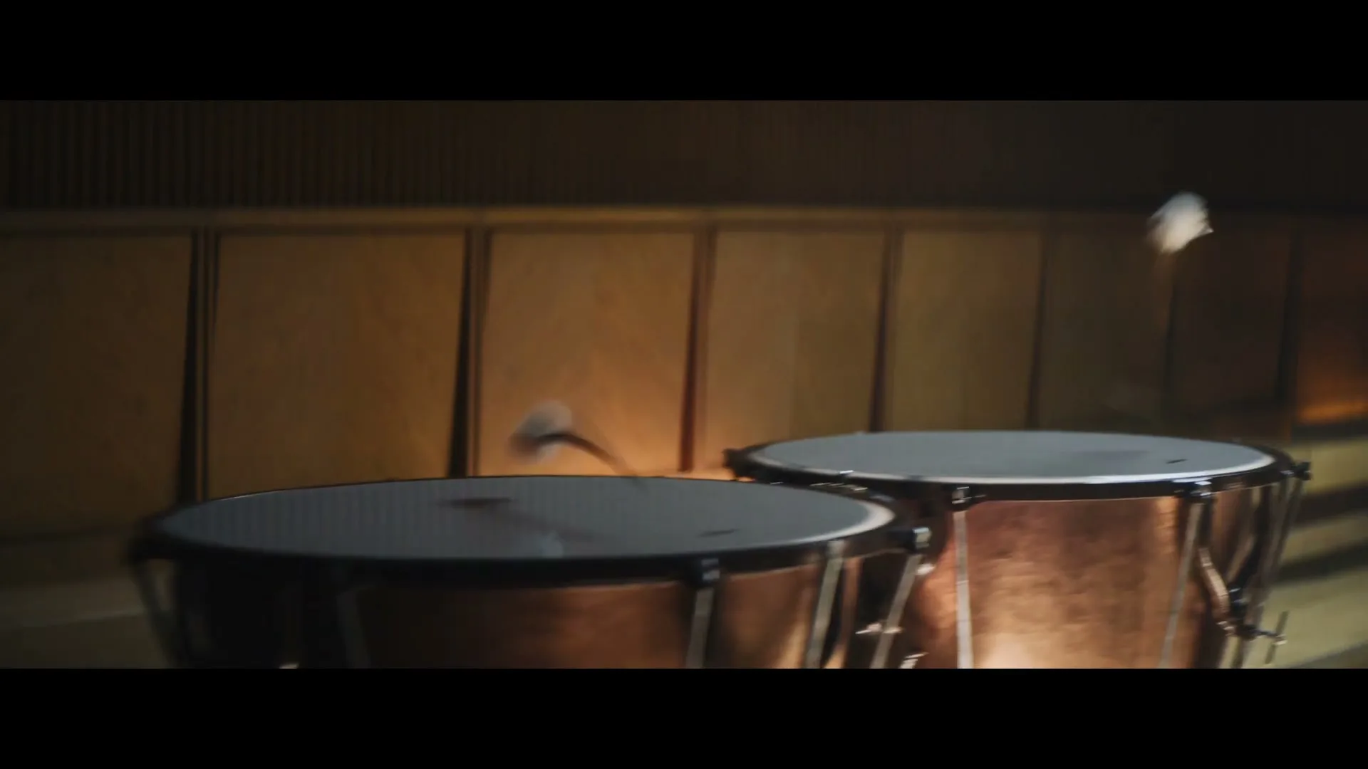 A pair of timpani drums are seen in a dimly lit orchestra pit, with drumsticks hitting them, creating a percussive sound.