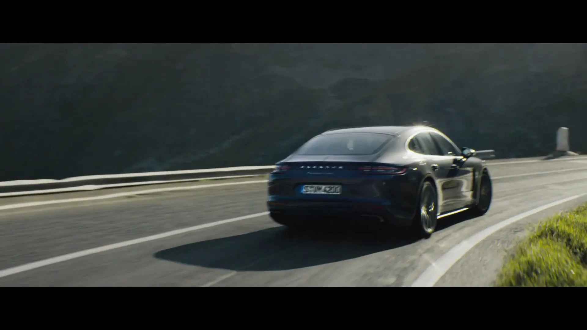 A dark grey Porsche Panamera Turbo drives away from the camera on a winding mountain road under a bright sky.