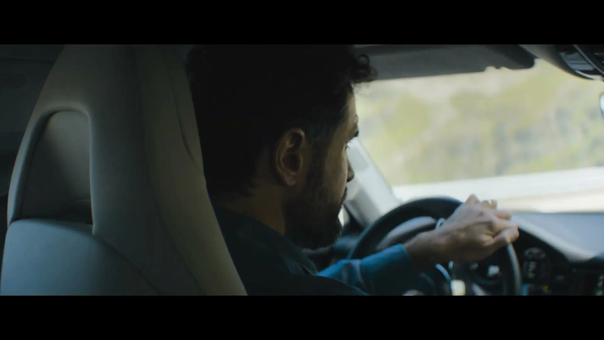 A man with dark hair and a beard drives a car, looking forward with a focused expression on a sunny day.
