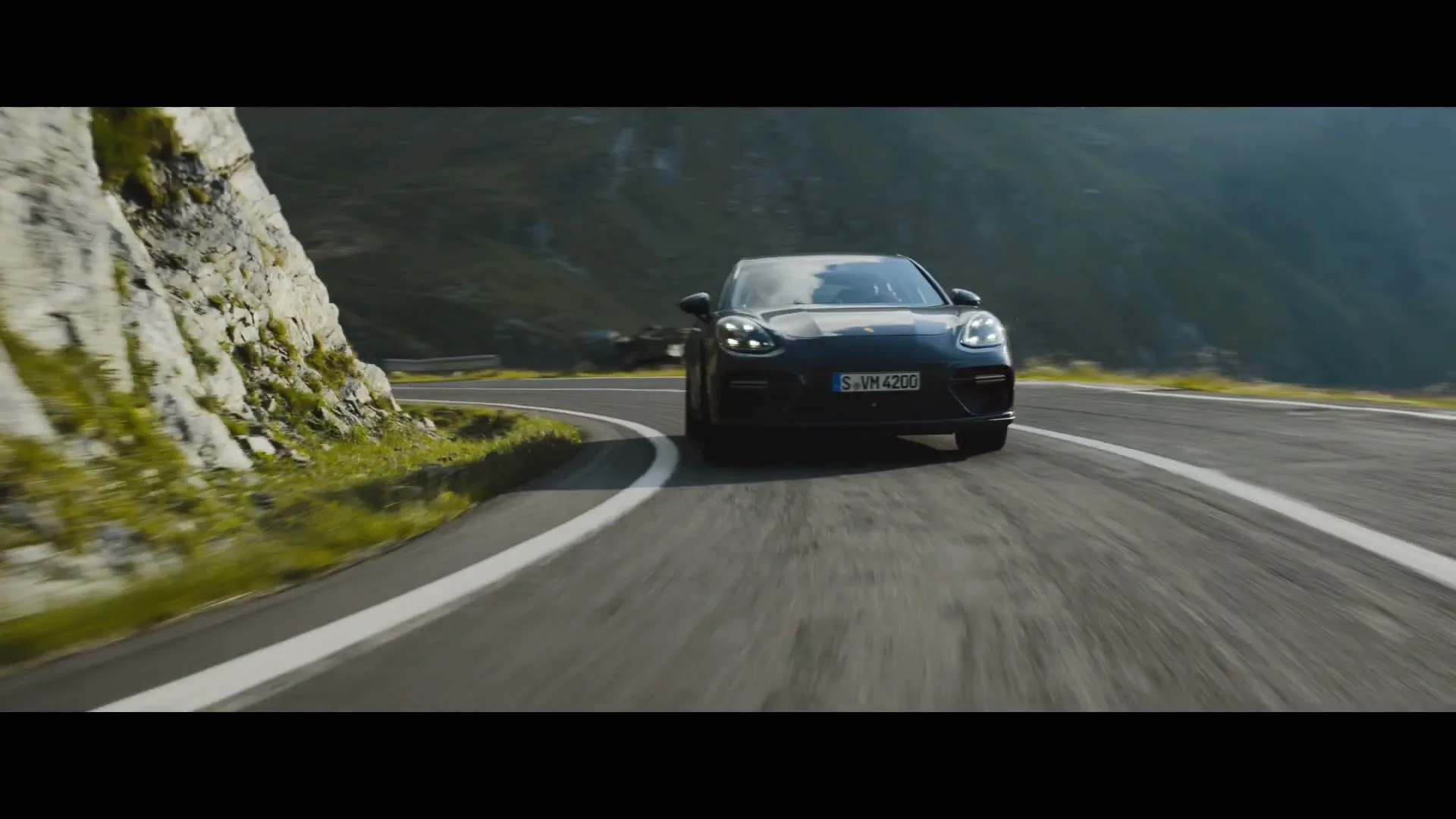 A dark grey Porsche Panamera drives quickly around a bend on a mountain road during the day.