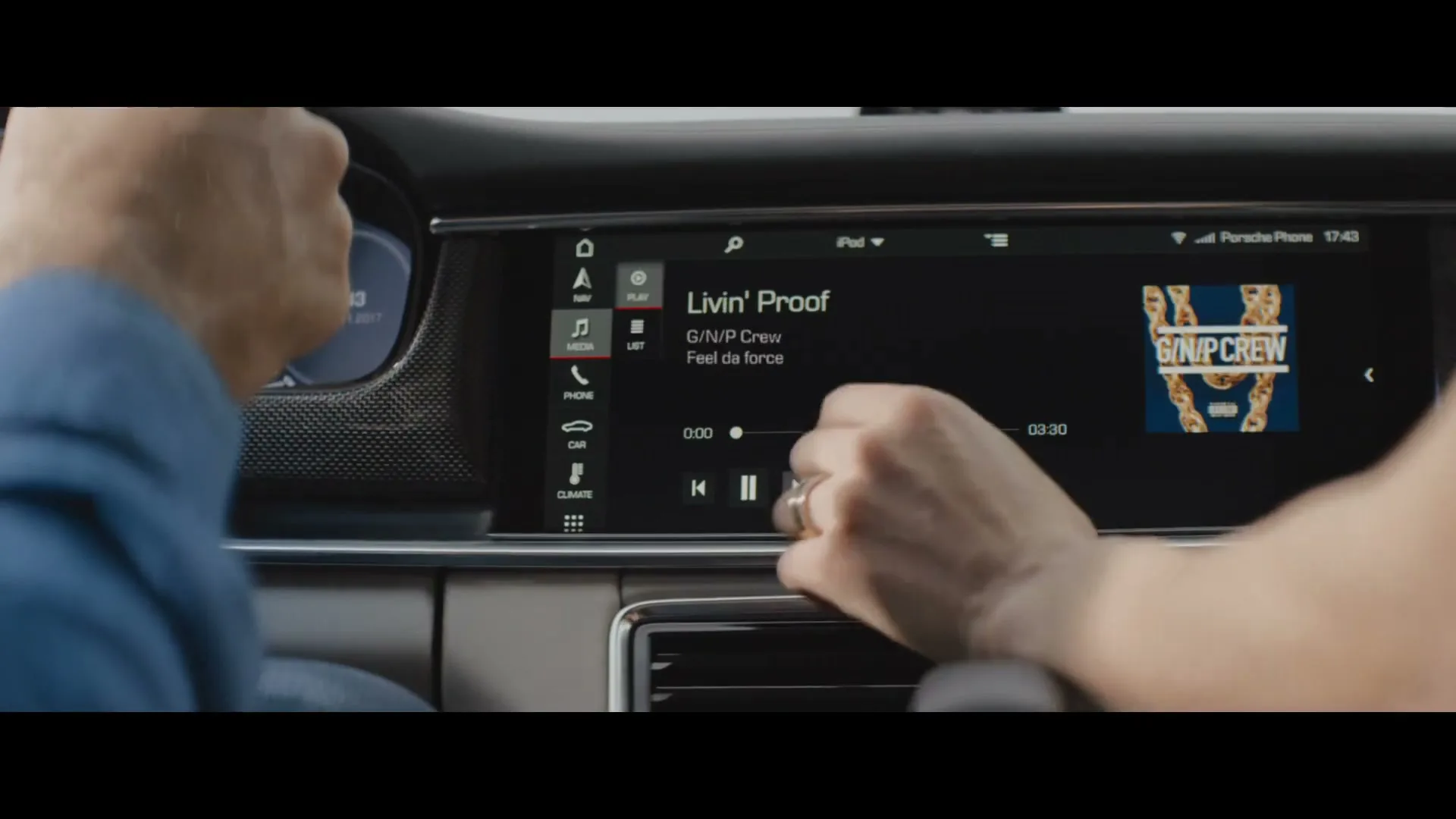 A man in a blue shirt drives a car while a woman's hand interacts with the car's infotainment system, changing music from classical to hip-hop.