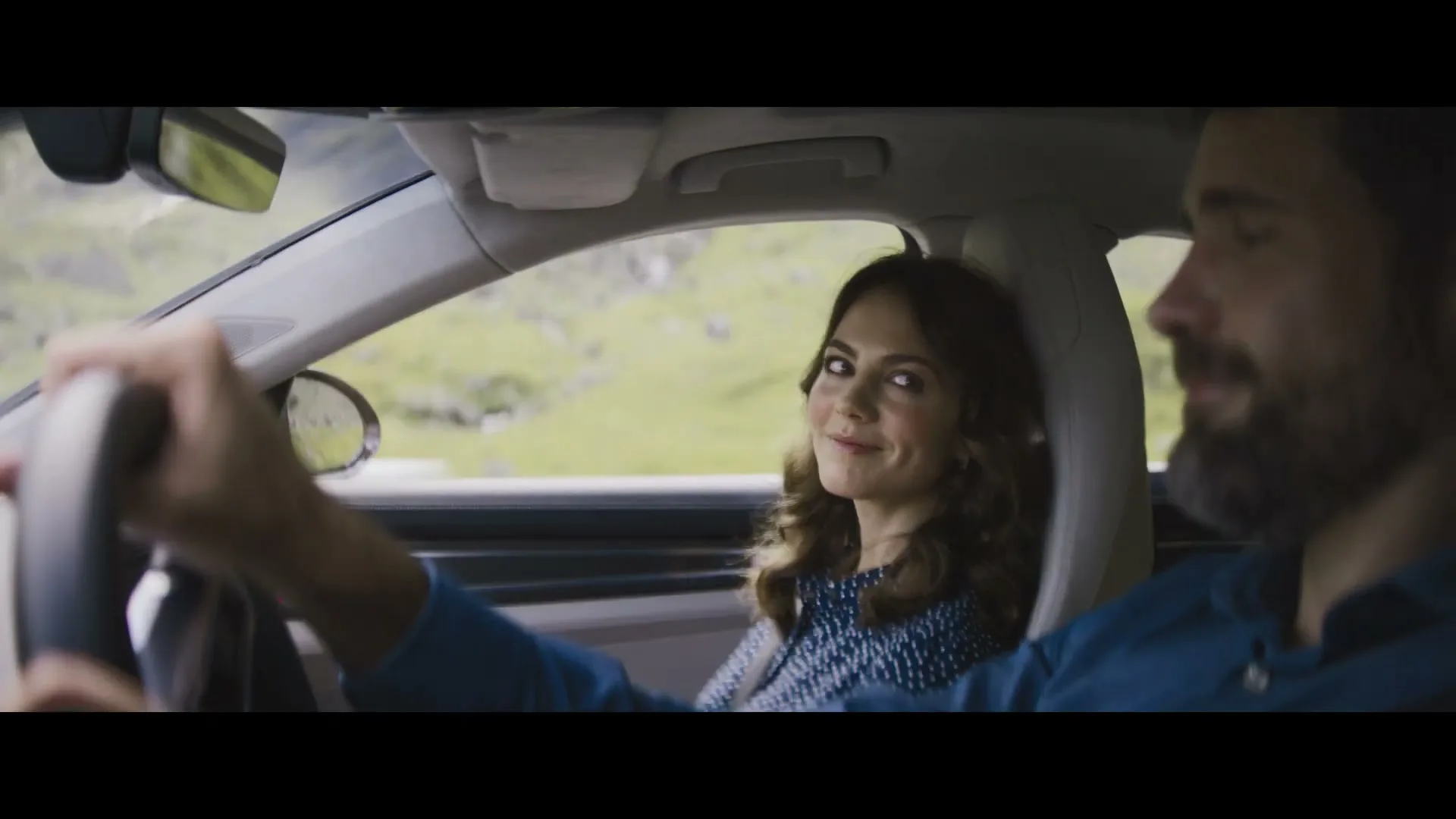 A woman with dark curly hair in a blue polka dot shirt smiles at the camera from the passenger seat of a car, while a man drives.