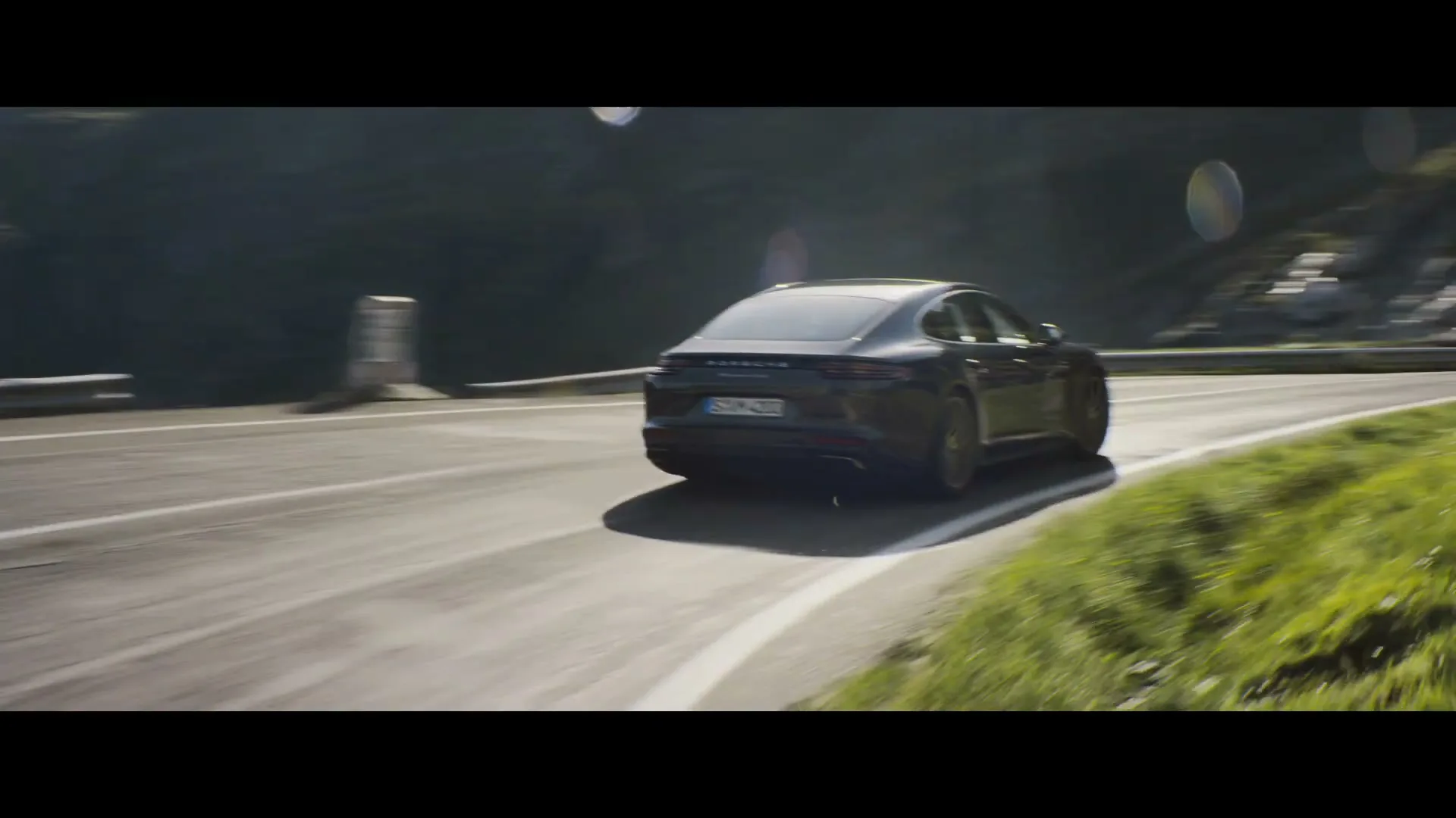 A dark grey Porsche Panamera drives quickly along a winding mountain road on a sunny day.