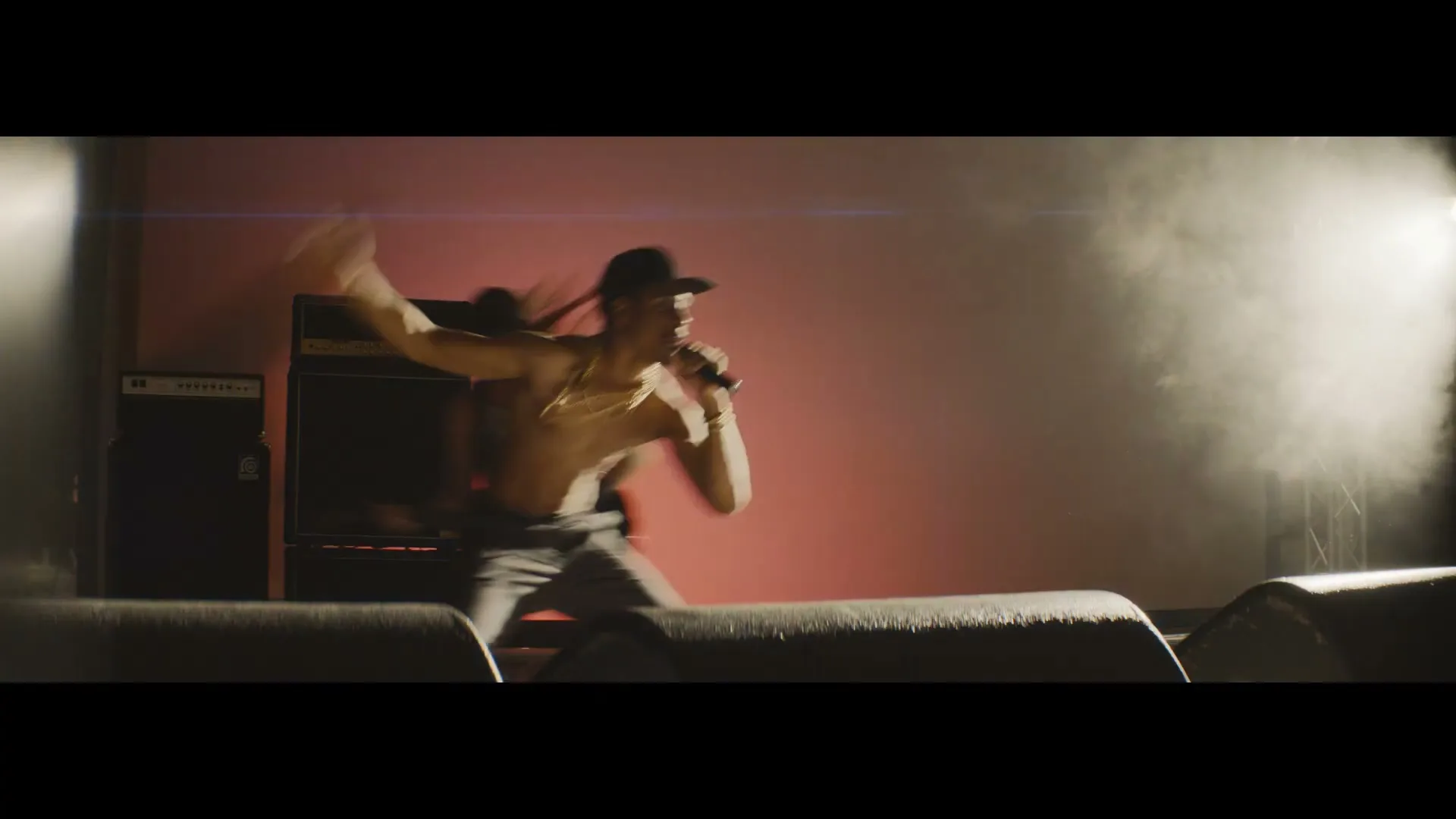 A male rapper, shirtless with a hat and chains, performs energetically on stage with a microphone, bathed in dramatic stage lighting.
