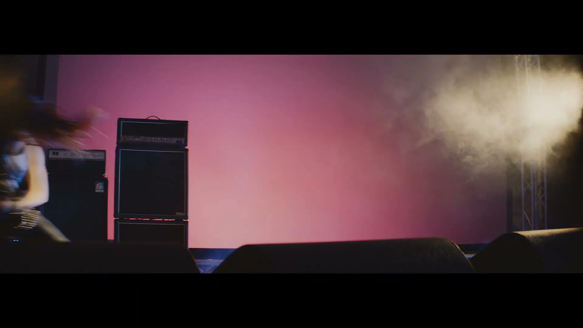 A female guitarist with long hair headbangs on a stage with amplifiers and smoke, illuminated by stage lights.