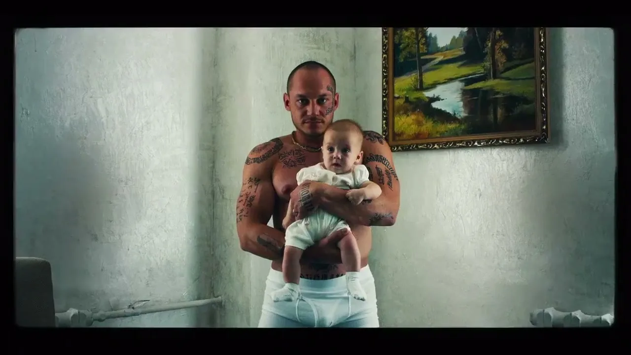 A heavily tattooed man with a shaved head, wearing only white briefs, holds a baby in a stark, white room with a landscape painting on the wall.