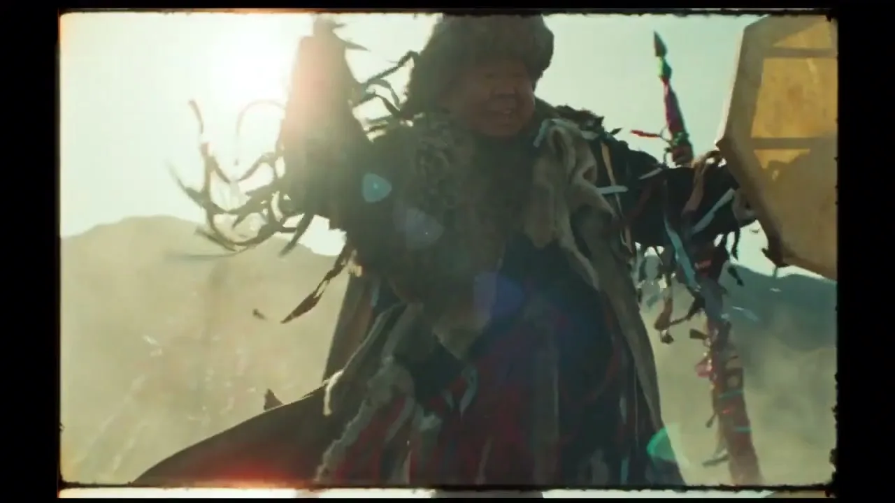A person in traditional shamanic attire with a fur hat and colorful ribbons performs a ritual, holding a drum in a bright, dusty outdoor setting.