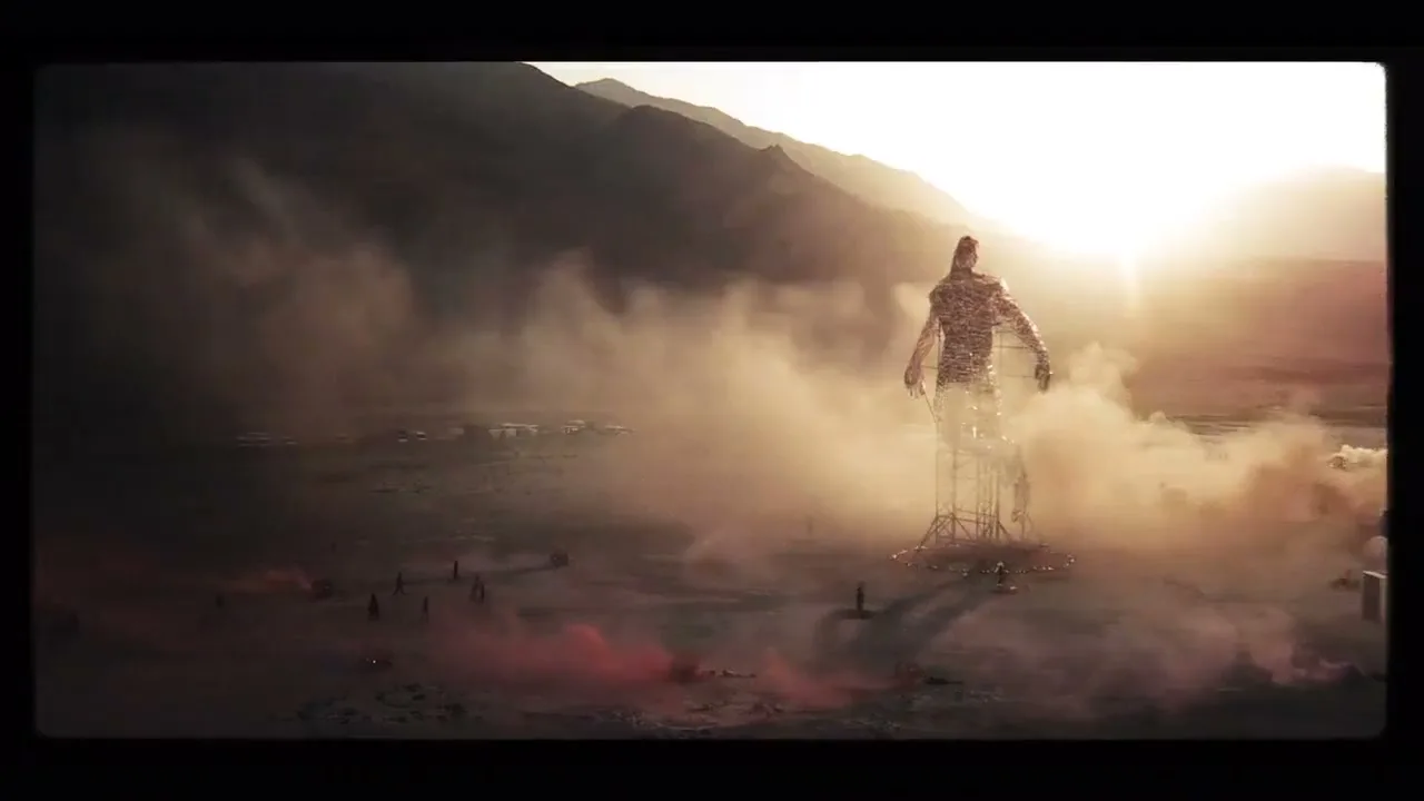 A giant, wire-frame human sculpture stands in a dusty desert landscape at sunset, surrounded by small figures and red smoke.