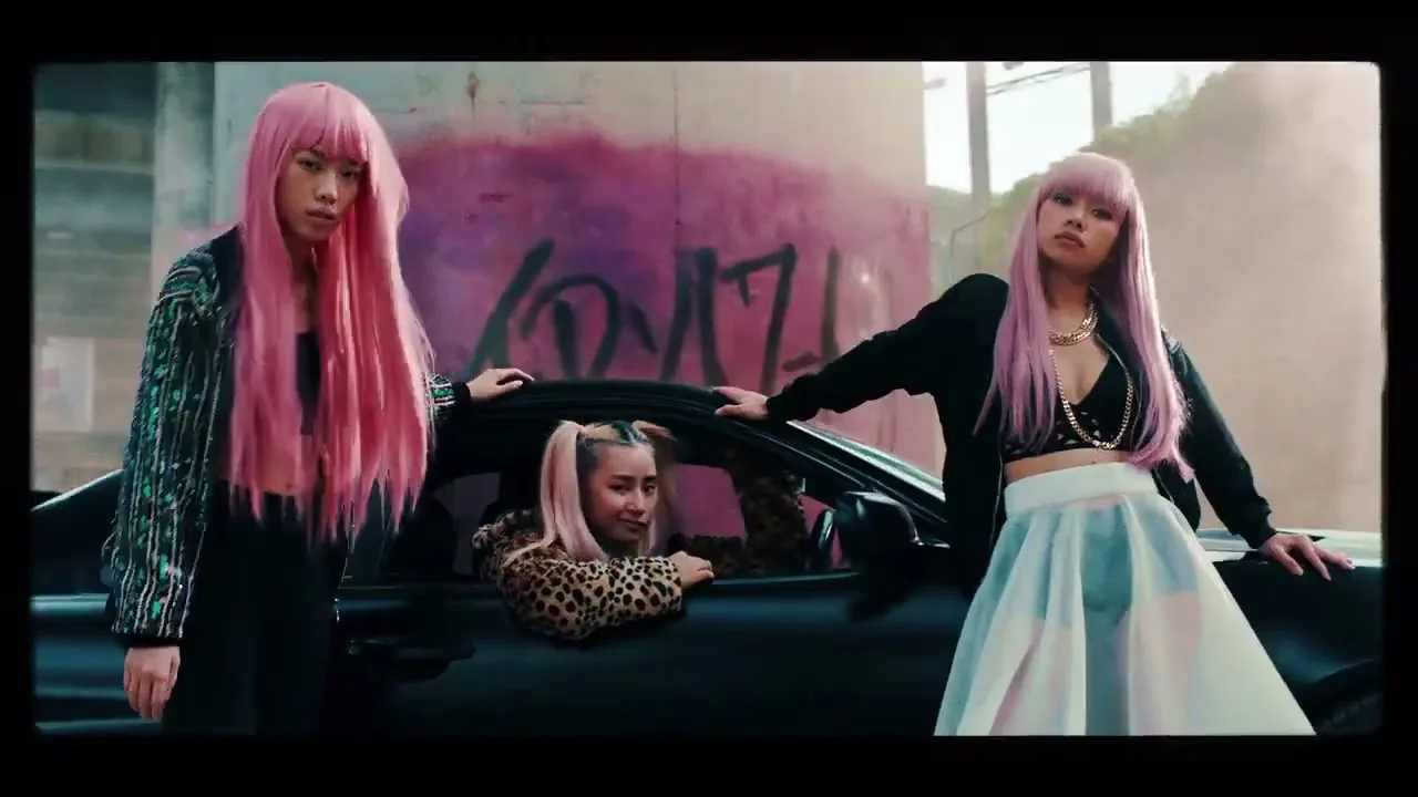 Three young Asian women with pink hair pose around a car in an urban setting with graffiti on a wall.
