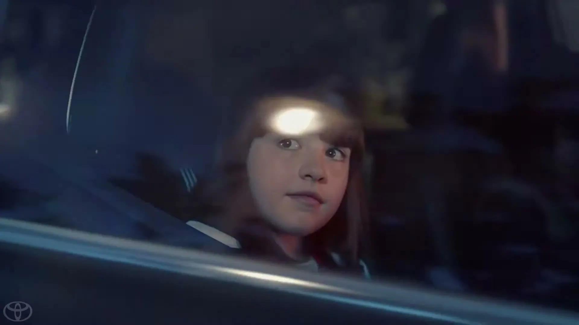 A young girl with dark hair and bangs looks out the car window with wide eyes, as if seeing something strange, at night.