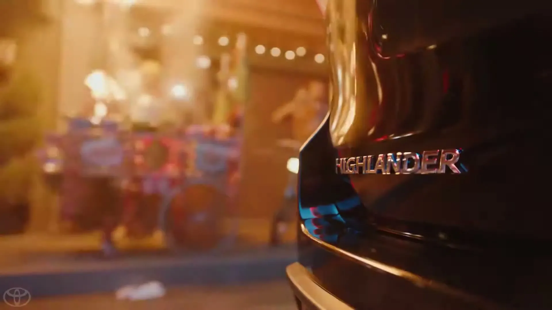 The rear of a black Toyota Highlander is visible on the right, with the "Highlander" emblem in focus, while a colorful street scene with blurred lights and a person on a bicycle is in the background.