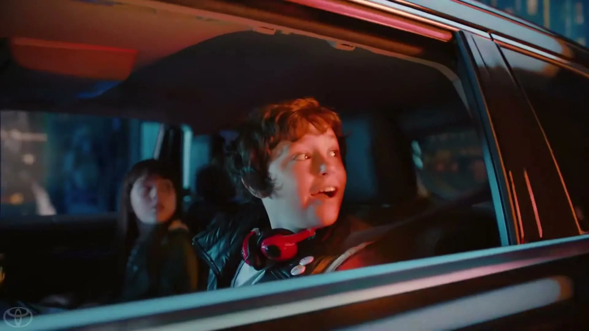 A young boy with curly hair, wearing headphones, looks out the car window with an excited expression while driving at night, with a girl in the back seat.
