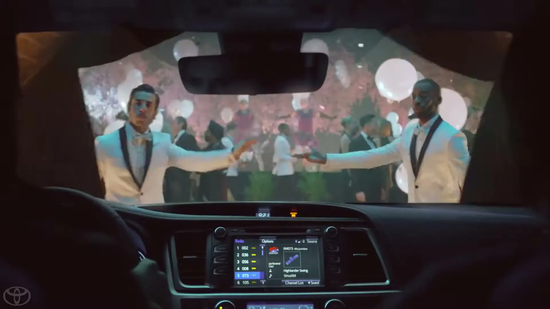 A driver's POV from inside a car, looking through the windshield at a party with people in white tuxedos and balloons.