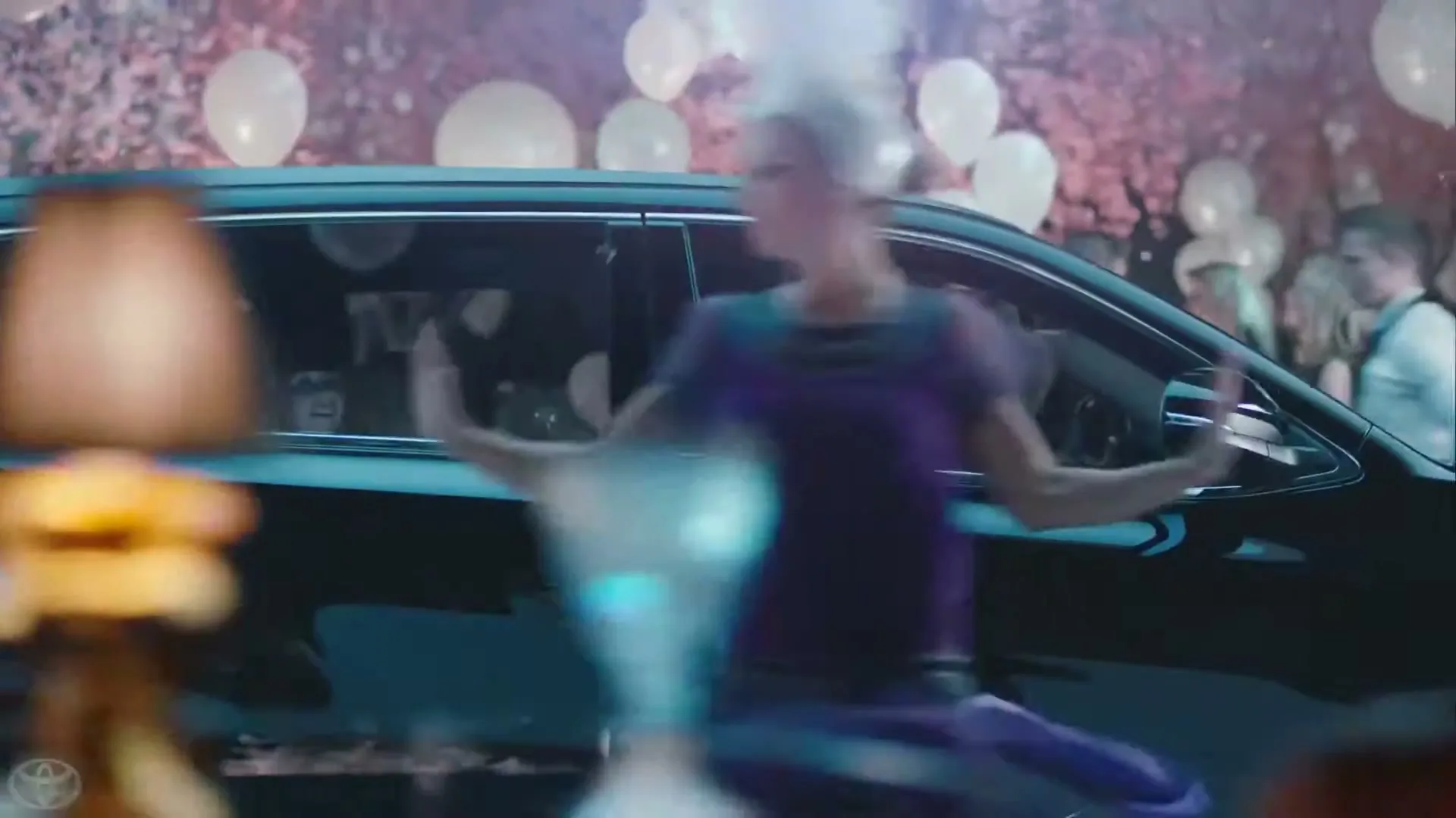 A black Toyota car drives past a crowd of people dancing at a party with balloons and pink foliage in the background.