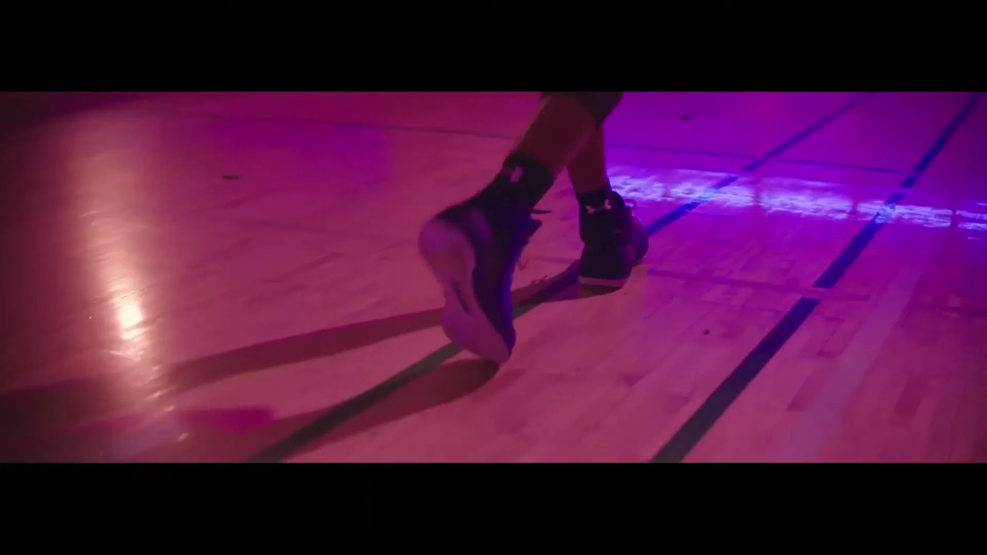 A person wearing black basketball shoes with pink soles walks across a wooden basketball court under purple and pink lighting.