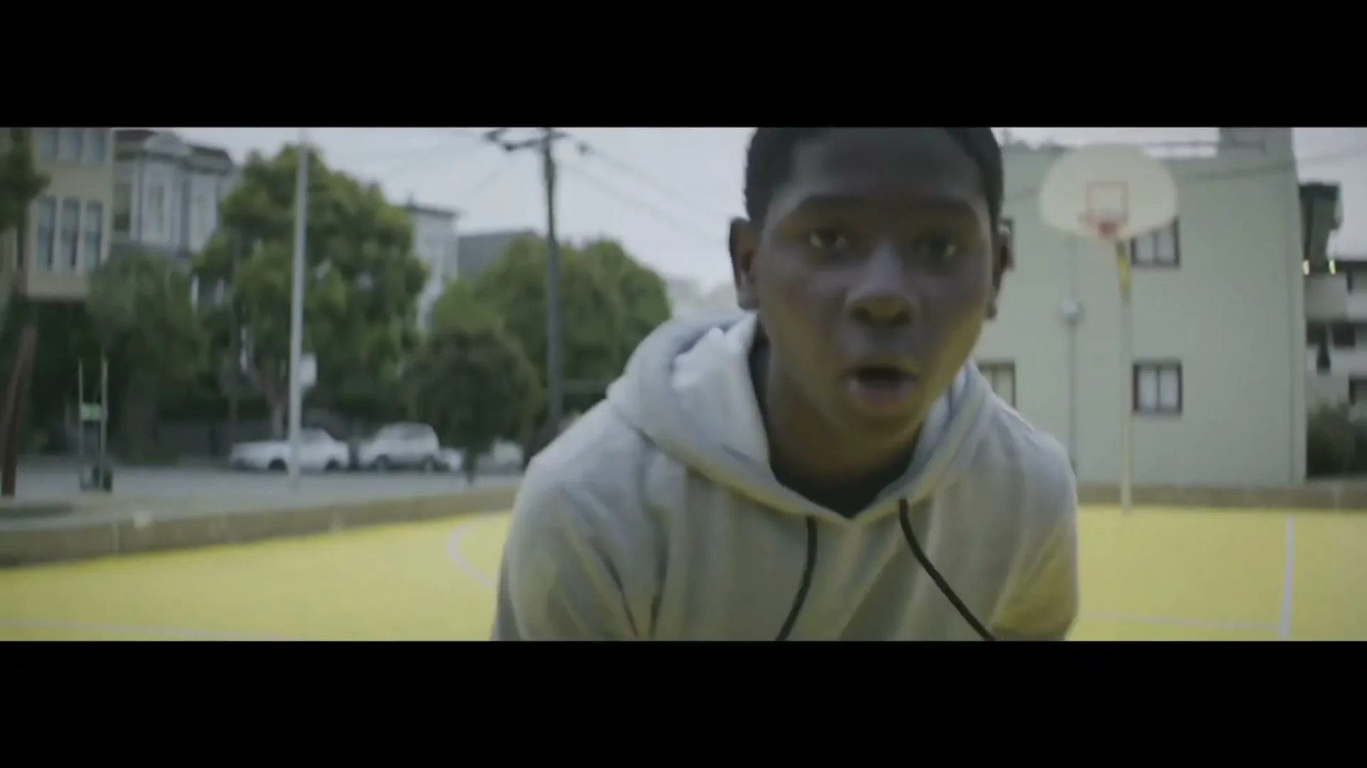 A young Black male in a grey hoodie dribbles a basketball on a yellow court in an urban neighborhood during the day.