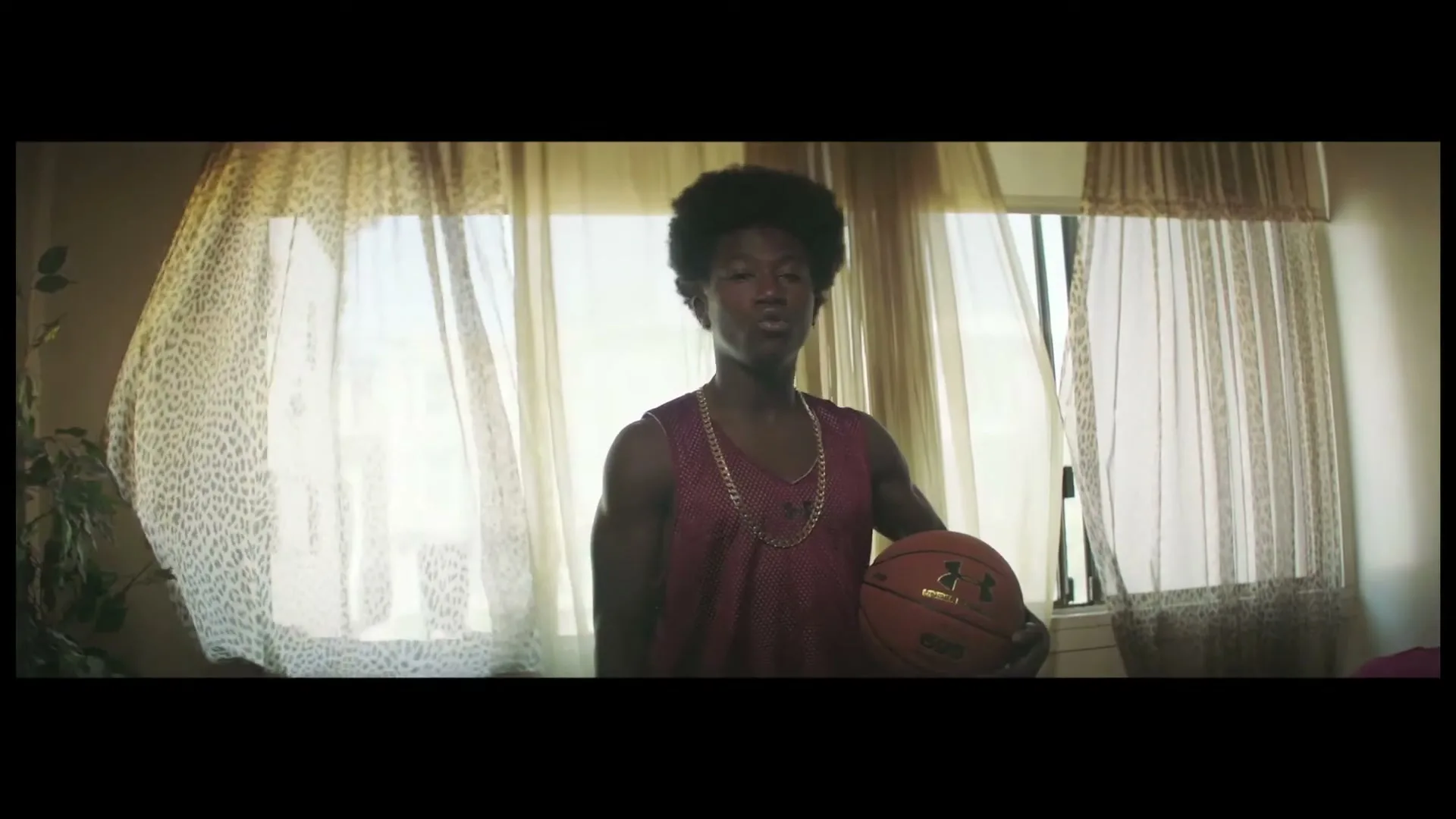 A young Black male with an afro, wearing a maroon basketball jersey and a gold chain, holds a basketball while standing in front of a window.