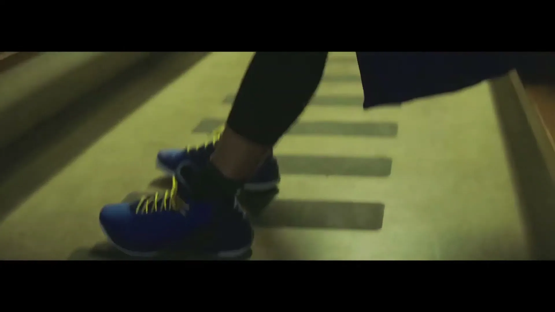 A person's feet in blue and yellow athletic shoes quickly step on a ladder agility drill on a green floor in an interior setting.