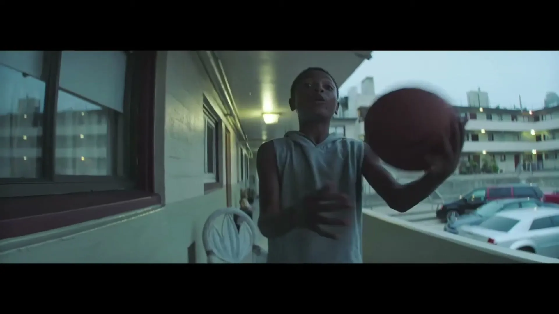 A young boy in a grey tank top dribbles a basketball on a balcony, with an apartment complex and parked cars in the background.