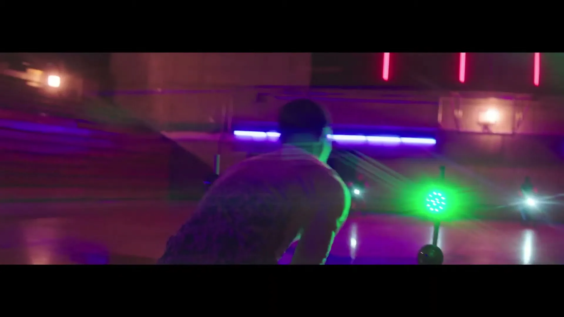 A male athlete in a dark tank top and cap is seen from behind, running on a track with bright green and purple lights.