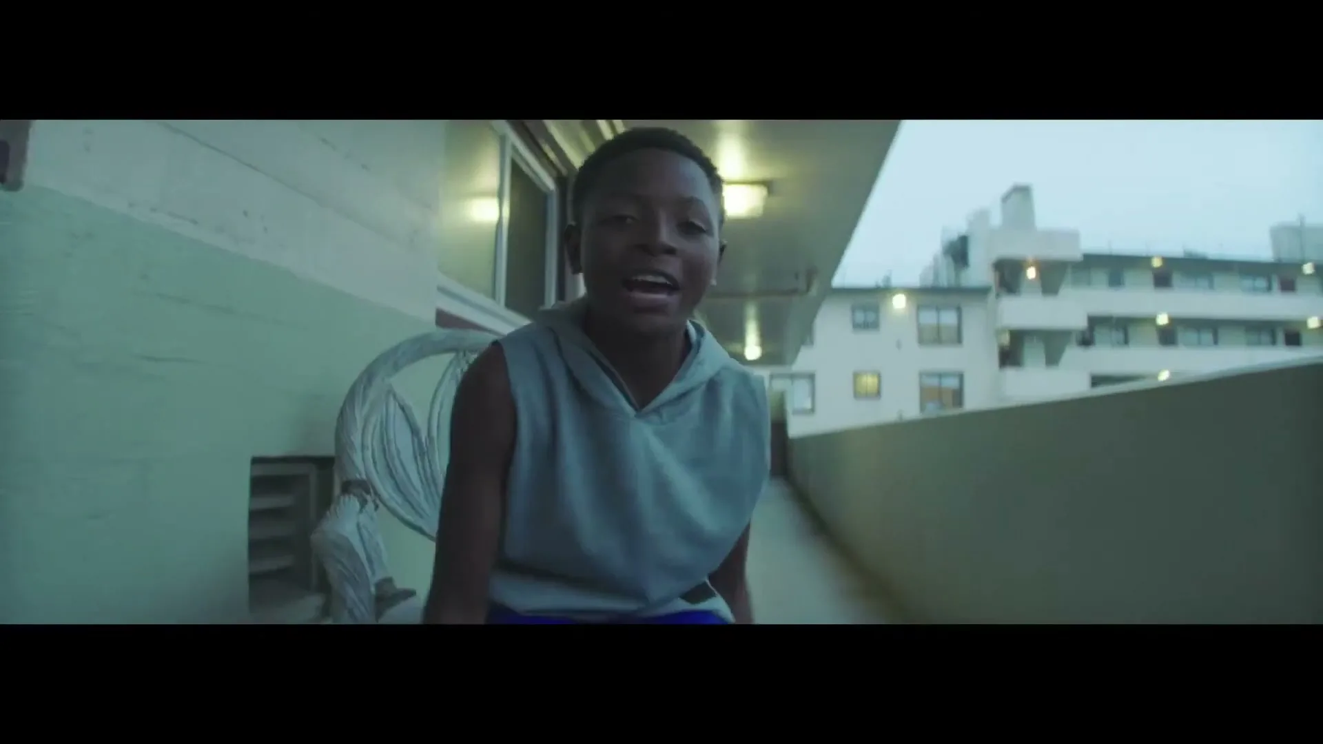 A young Black boy in a grey sleeveless hoodie looks directly at the camera, speaking intensely on an outdoor balcony at dusk.