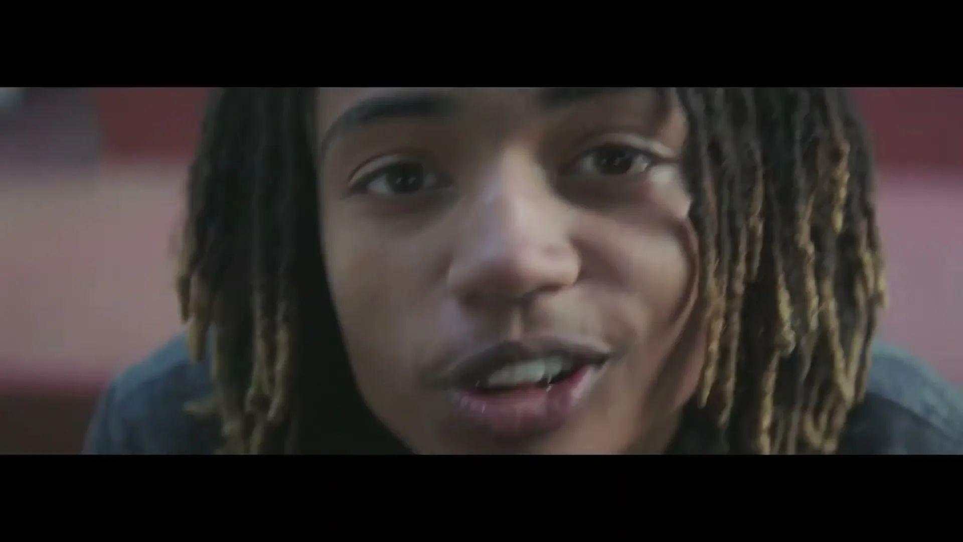 A young male with dreadlocks looks directly at the camera, his mouth slightly open as if speaking or reacting, in an interior setting.