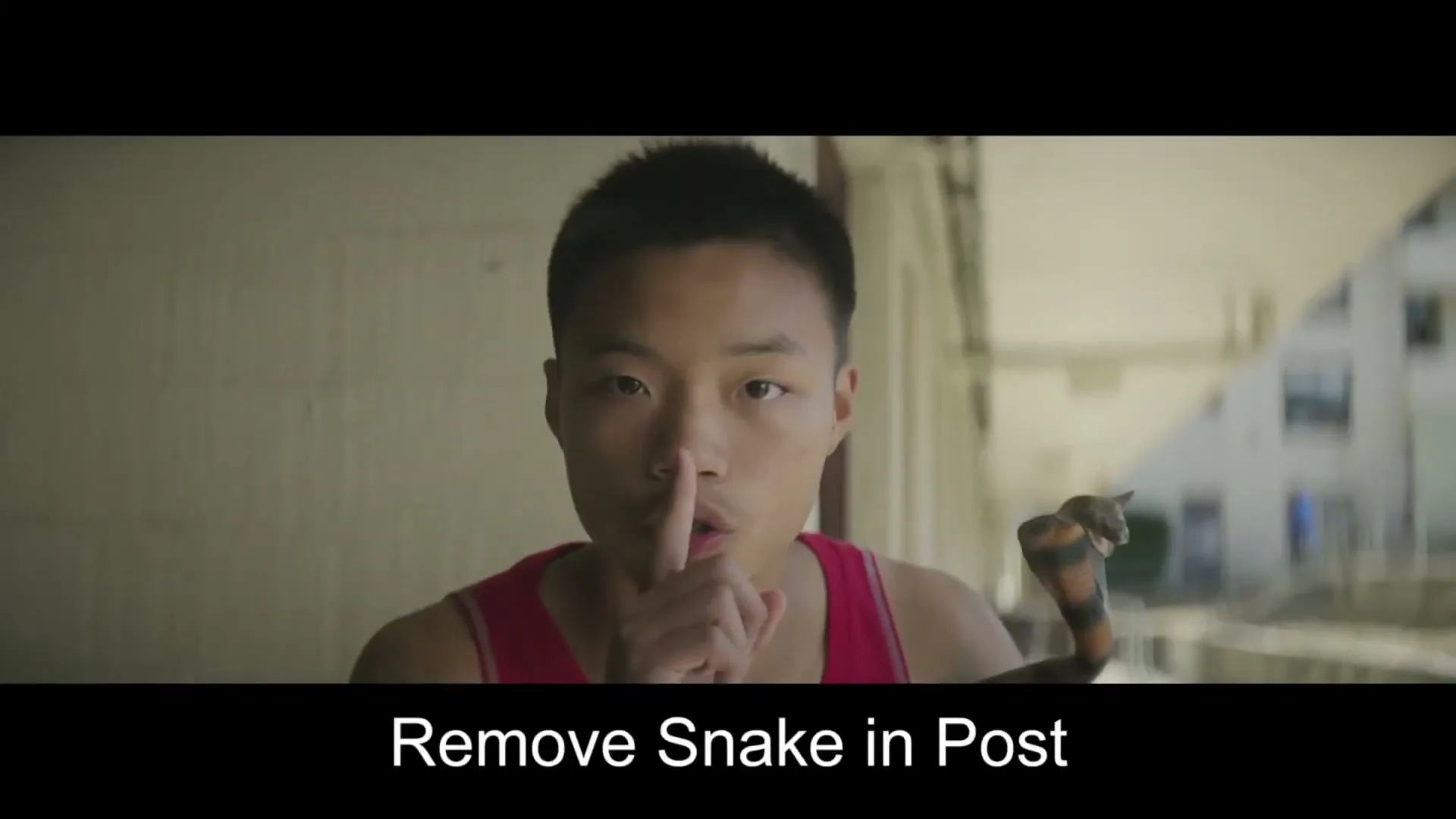 A young male basketball player in a red tank top holds a finger to his lips, shushing the viewer, with a snake on his shoulder in an outdoor urban setting.
