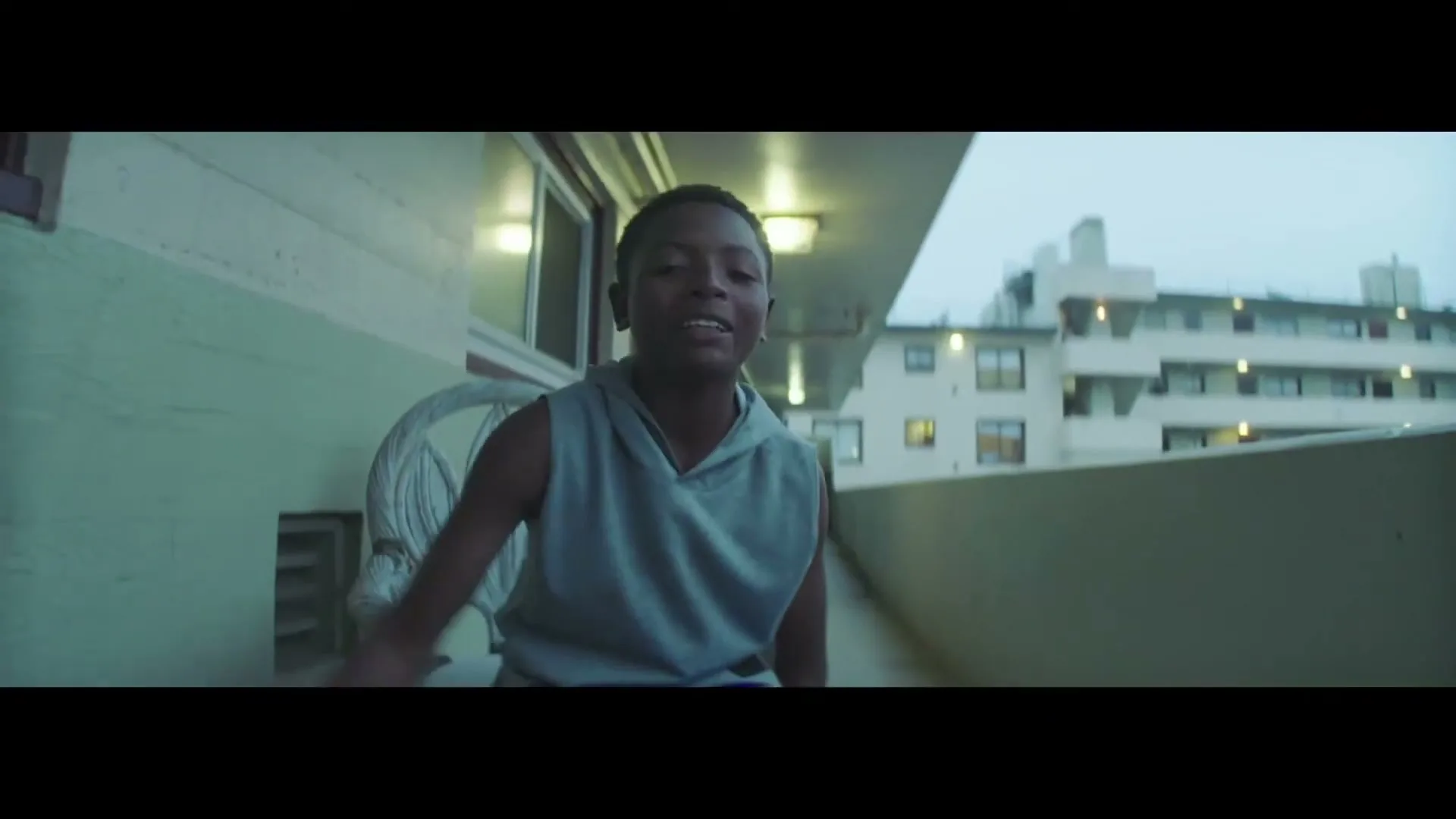 A young boy in a grey sleeveless hoodie sits on a balcony, looking directly at the camera and speaking.
