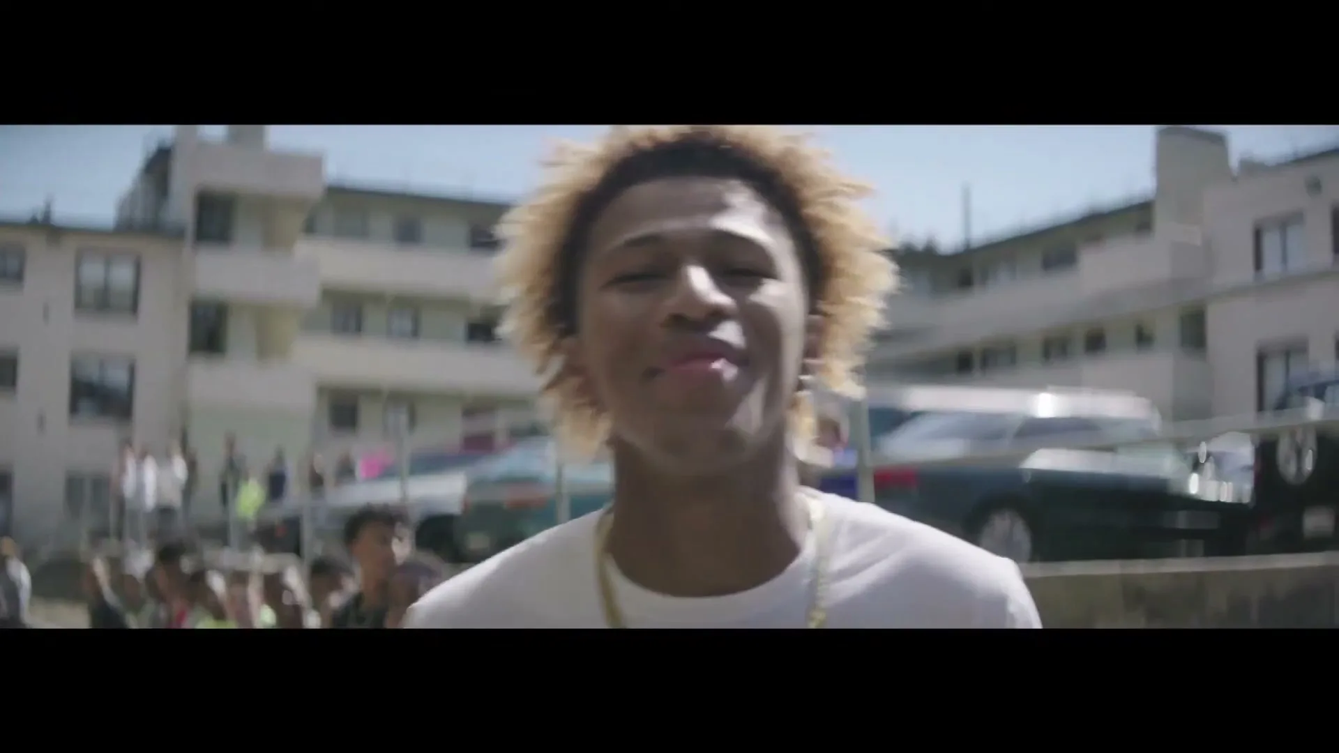 A young male with curly blonde hair and a white t-shirt smiles at the camera in an outdoor urban setting with apartment buildings in the background.