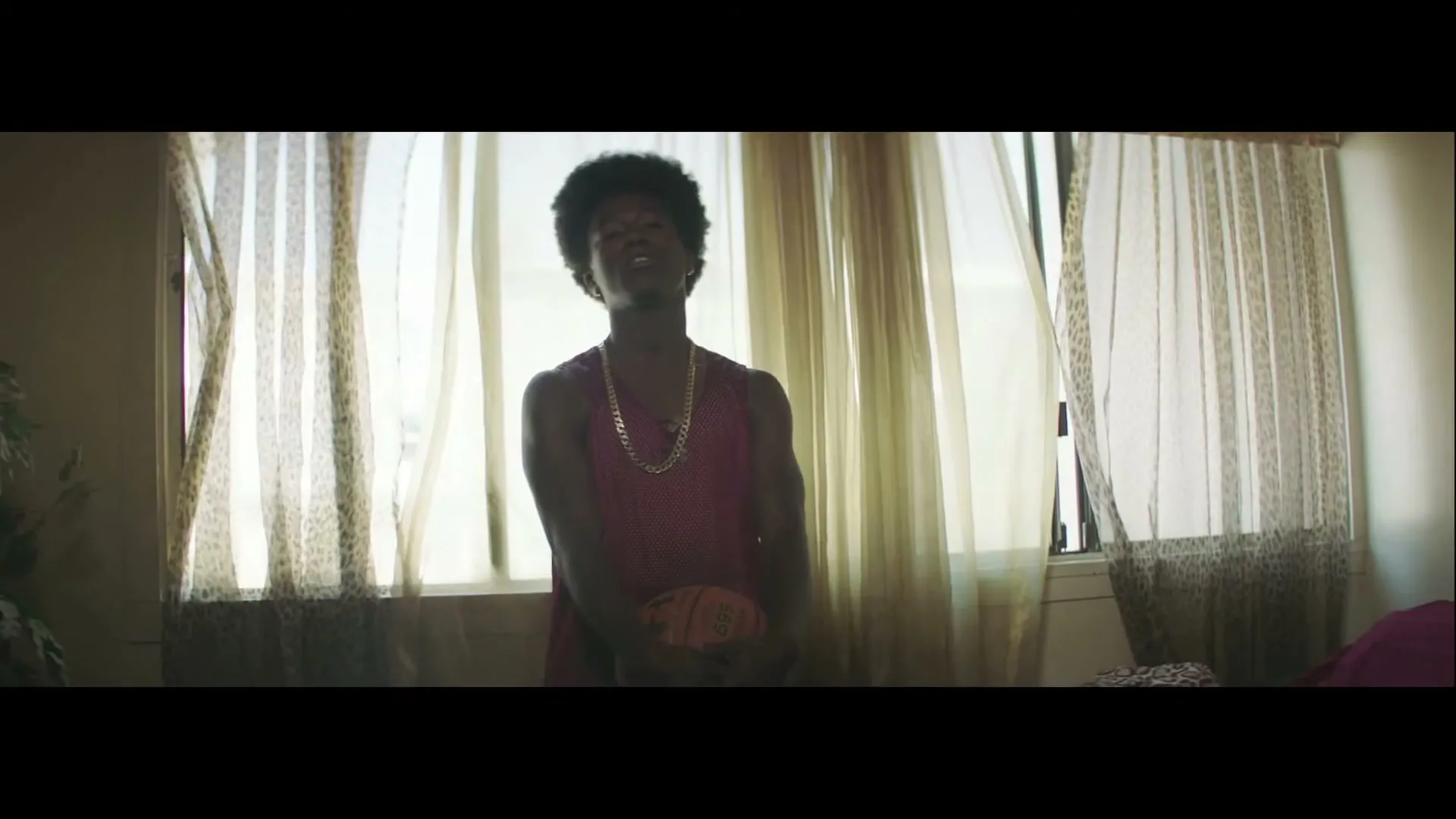A young man with an afro, wearing a pink mesh tank top and gold chain, holds a basketball in a sunlit room.