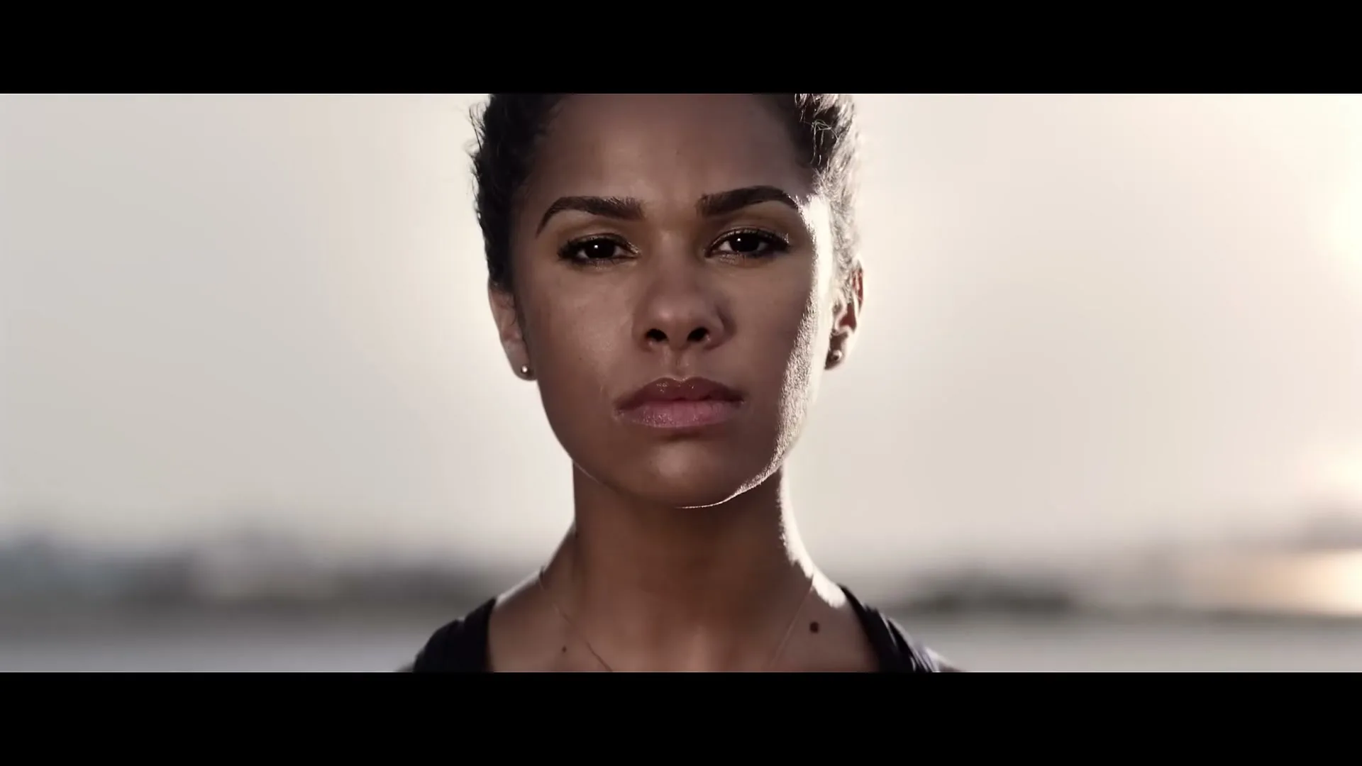 A determined female athlete with dark hair and a serious expression looks directly at the camera, outdoors at dusk.
