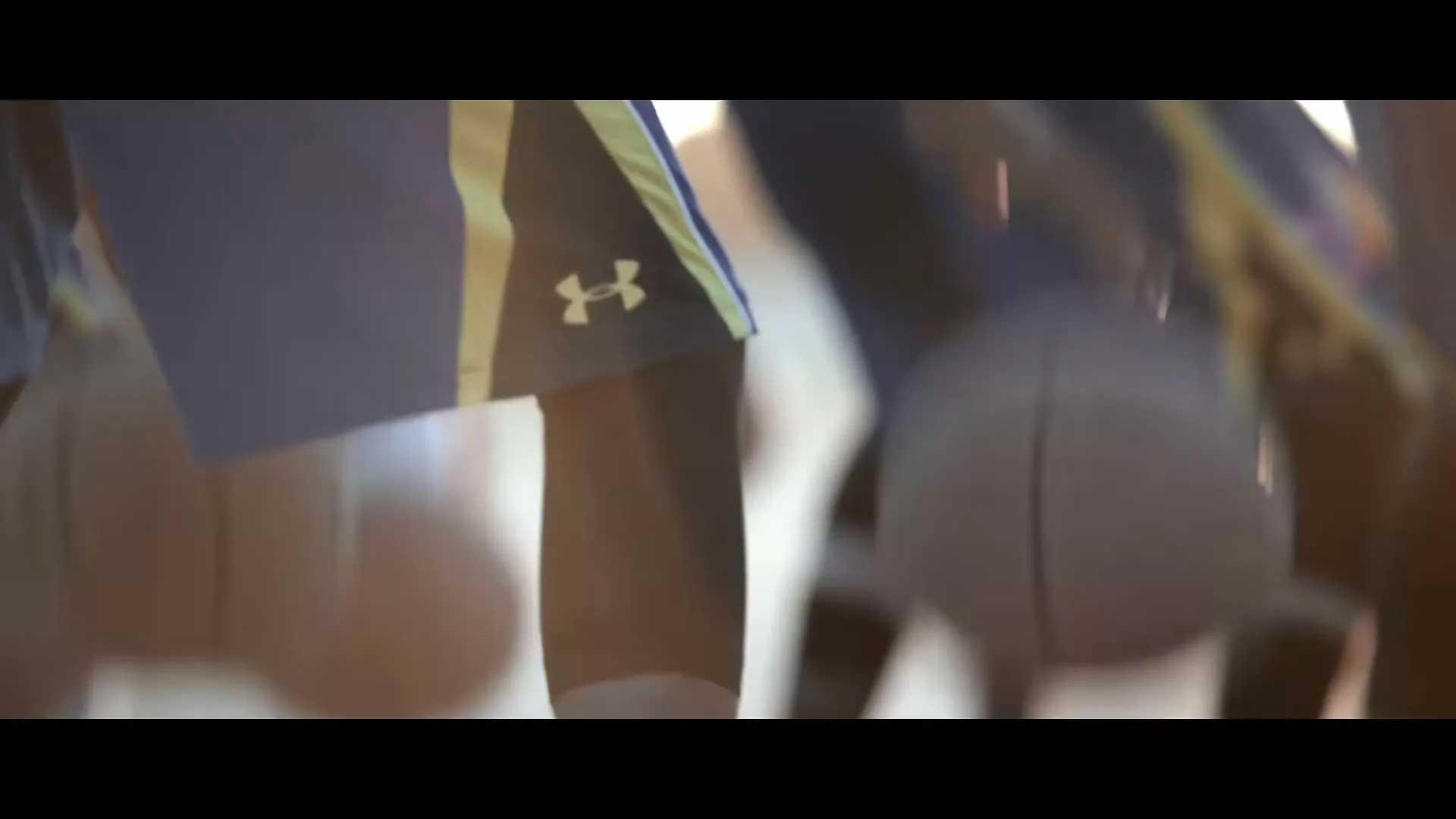 A basketball player in blue and yellow shorts with an Under Armour logo dribbles a basketball on a court, with other players blurred in the background.