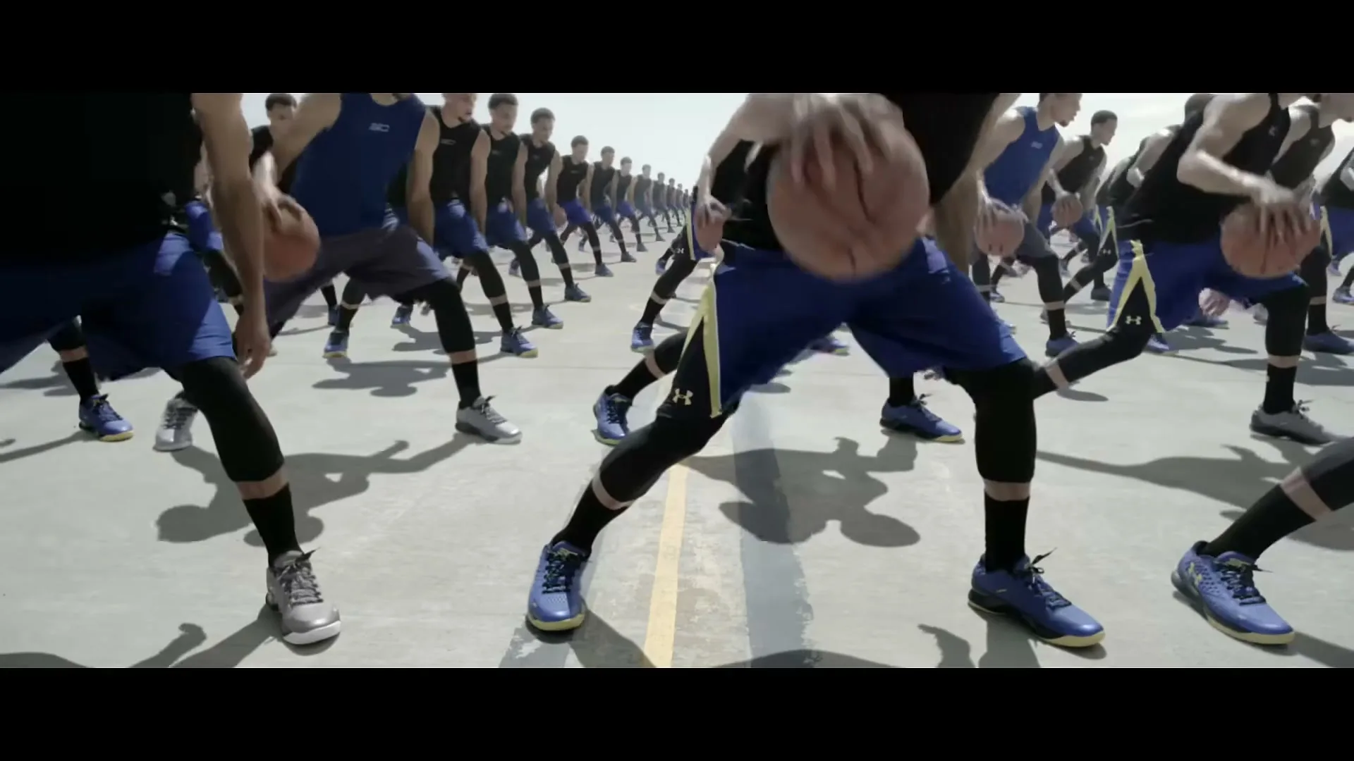 A group of male basketball players in blue and black uniforms dribble basketballs in unison on a concrete court under a bright sky.