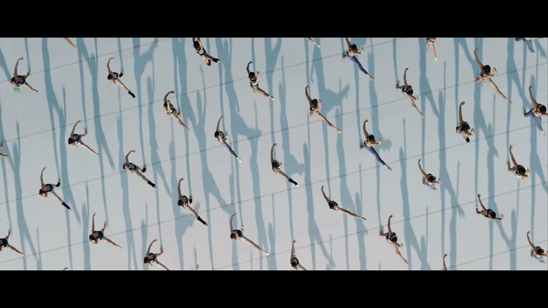 A top-down view shows many female gymnasts in black leotards performing synchronized movements on a light blue floor with shadows.