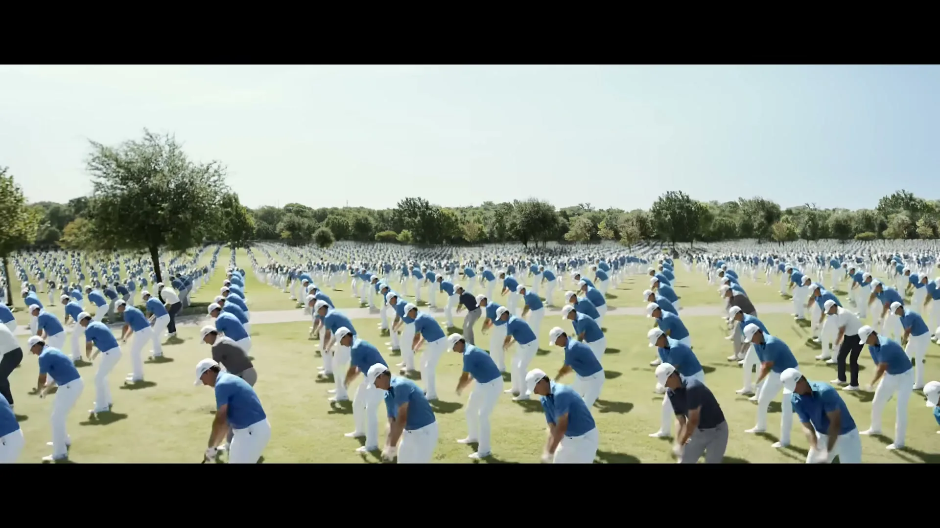 Hundreds of male golfers in blue shirts and white pants swing golf clubs in unison on a vast green golf course under a clear sky.