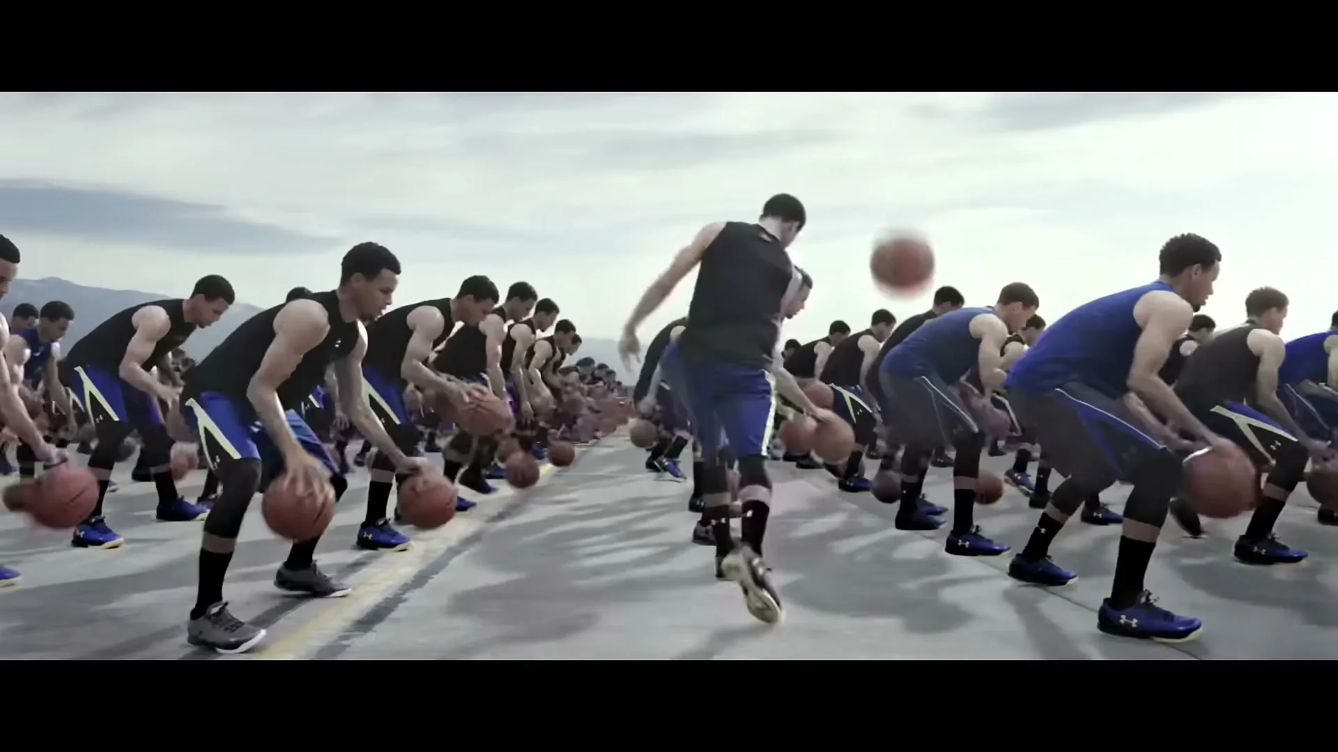Dozens of identical Stephen Curry clones in Under Armour gear dribble basketballs in unison on an outdoor court under a cloudy sky.