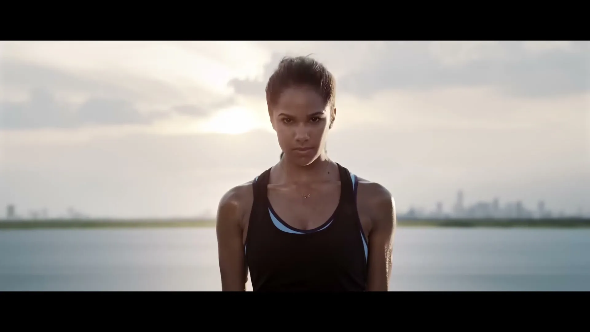 A female athlete in a black tank top looks up at the camera, with other athletes blurred behind her, outdoors by water at sunset.