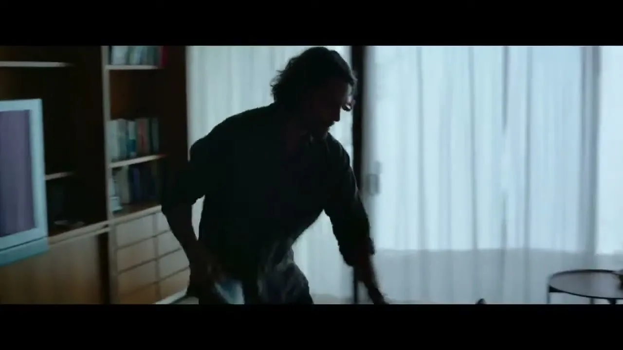 A man with dark, curly hair, wearing a dark shirt, walks across a modern living room, picking up items as he goes.