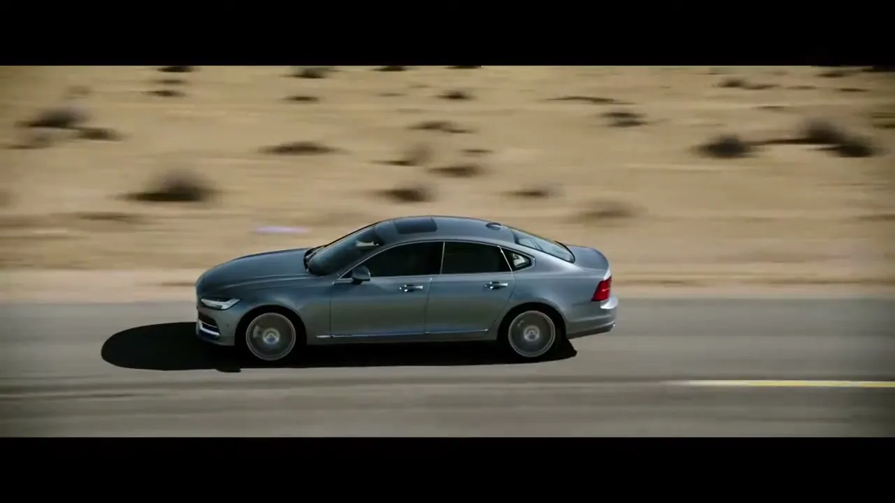 A grey Volvo S90 sedan drives quickly from left to right on a desert road under a bright sky, with blurred background.
