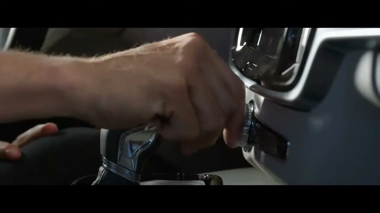 A man's hand is shown in a medium closeup, turning a knob to start a car in an interior setting.