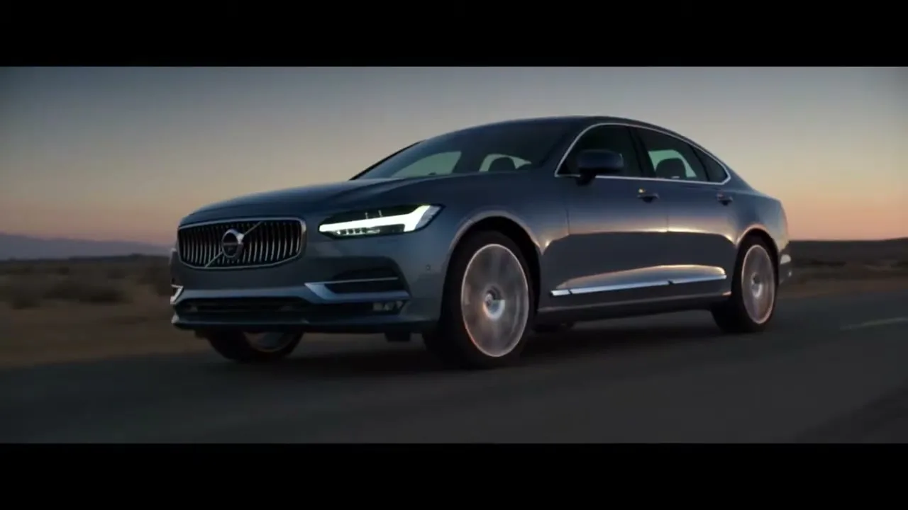 A dark grey Volvo S90 sedan drives on a desert road at dusk, with its headlights on, moving towards the right of the frame.