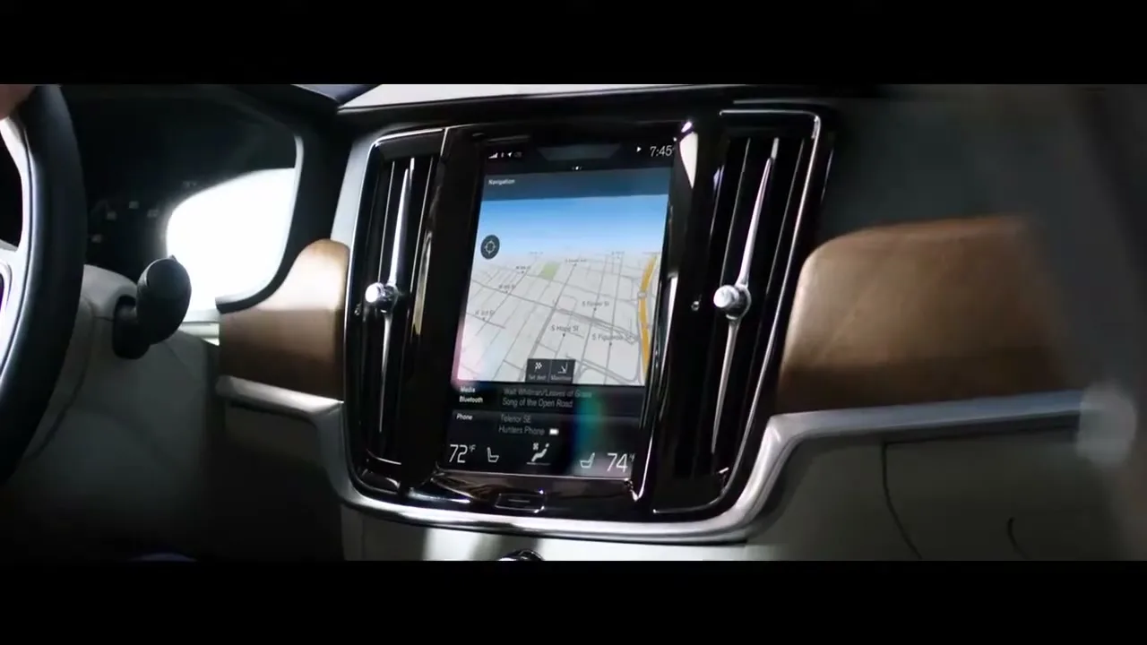 A close-up shot of a car's dashboard, showing the navigation screen with a map and climate controls, as a person's hand is visible on the steering wheel.