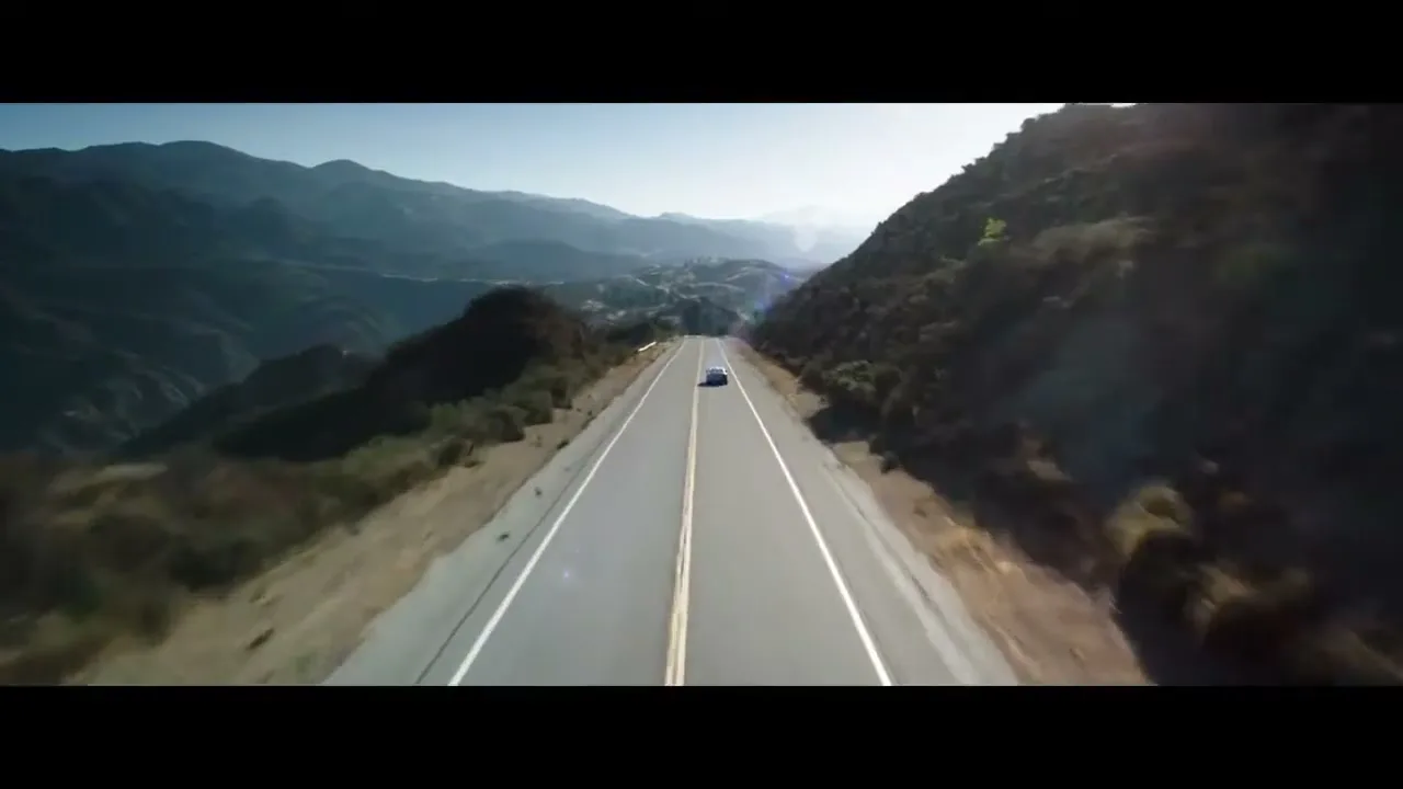 A blue Volvo S90 drives down a long, empty highway through a mountainous landscape under a clear sky.