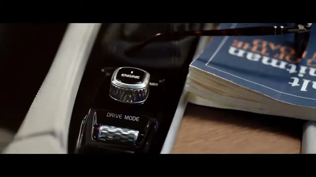 A close-up shot of a car's interior, showing the engine start/stop button and drive mode selector, with a book and glasses resting nearby.