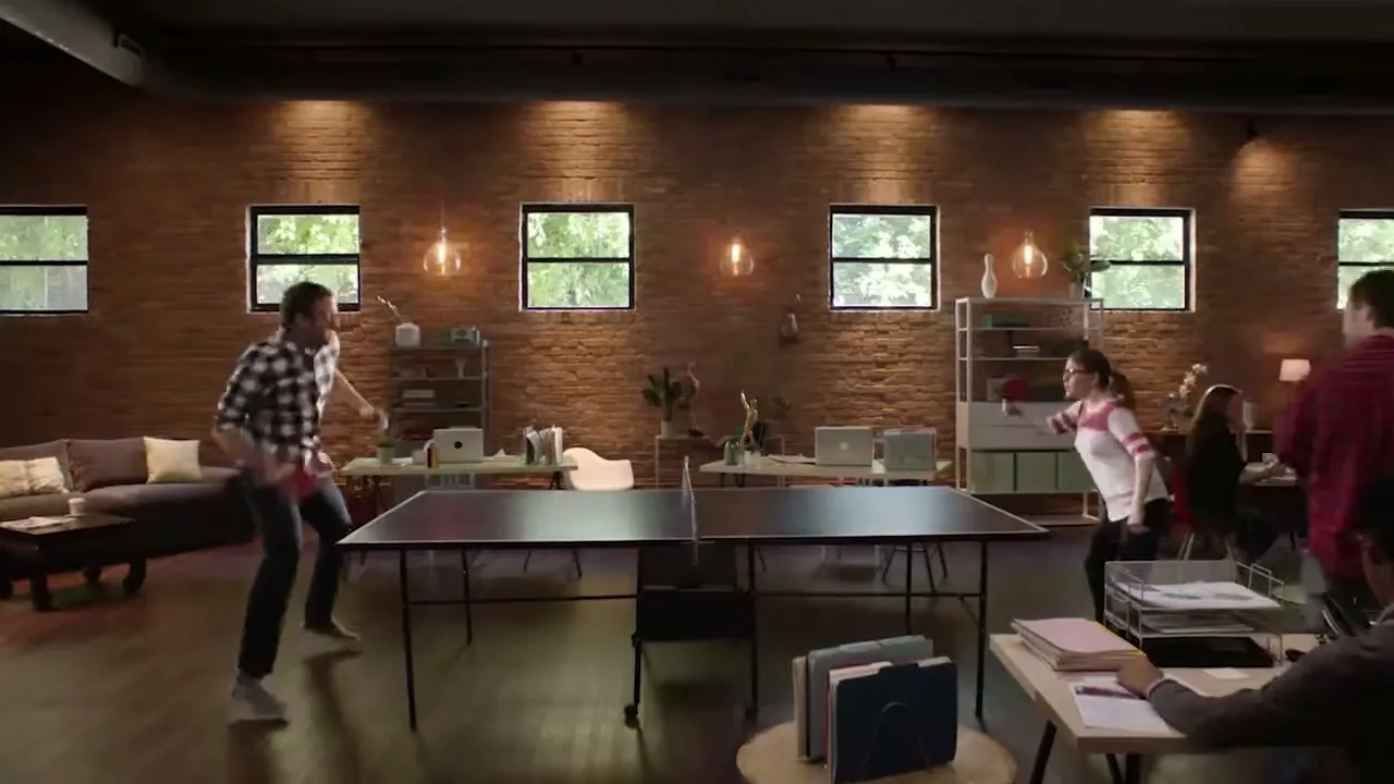 A man and a woman play ping pong in a modern office with exposed brick walls and large windows, while other colleagues work in the background.