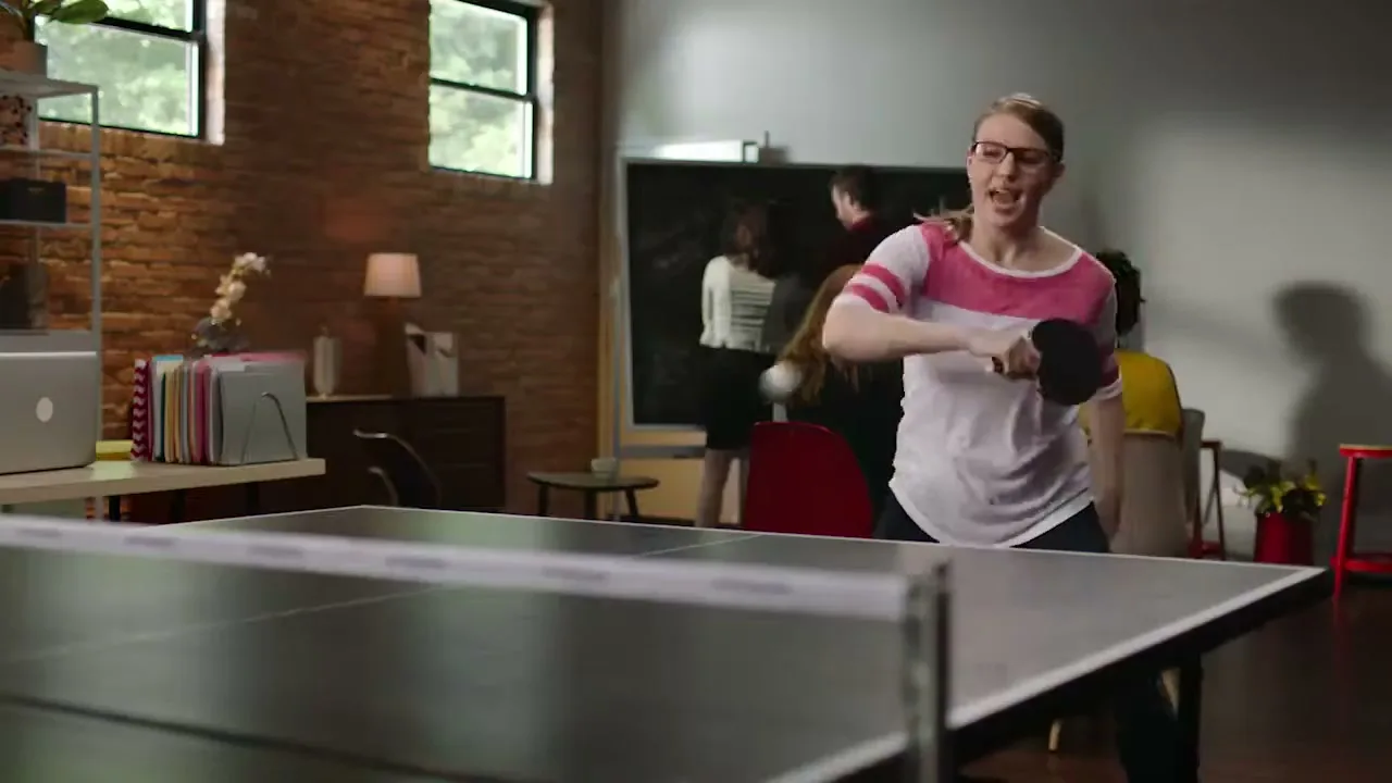 A young woman with glasses and a striped shirt plays ping pong in an office, with other people in the background.