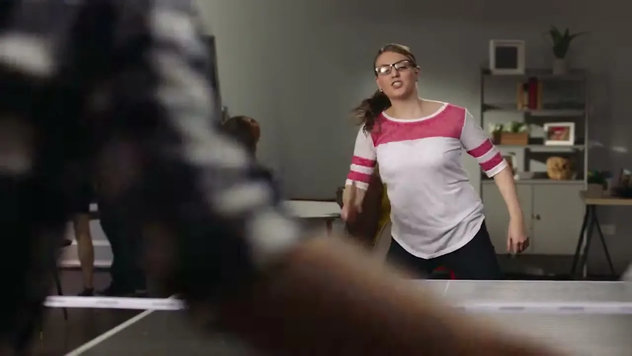 A woman in glasses and a striped shirt plays ping pong in an office, with other people in the background.