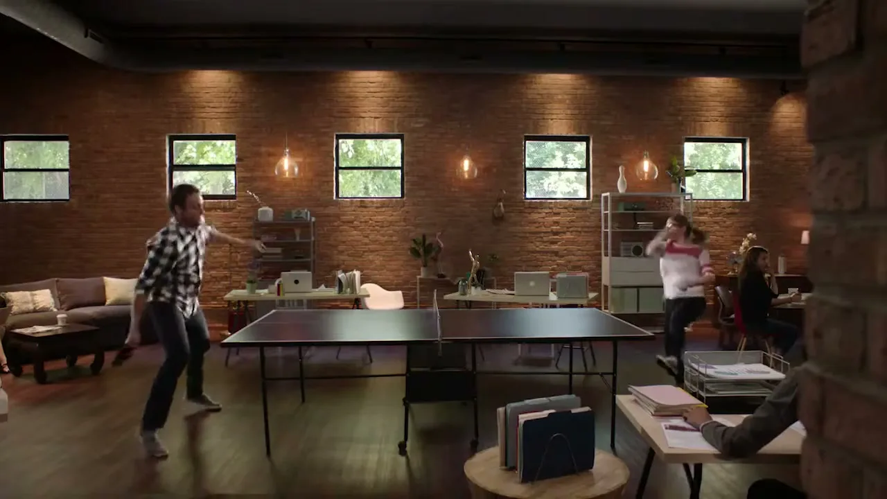 A man and a woman play ping pong in a modern office with exposed brick walls and large windows, while other employees work in the background.
