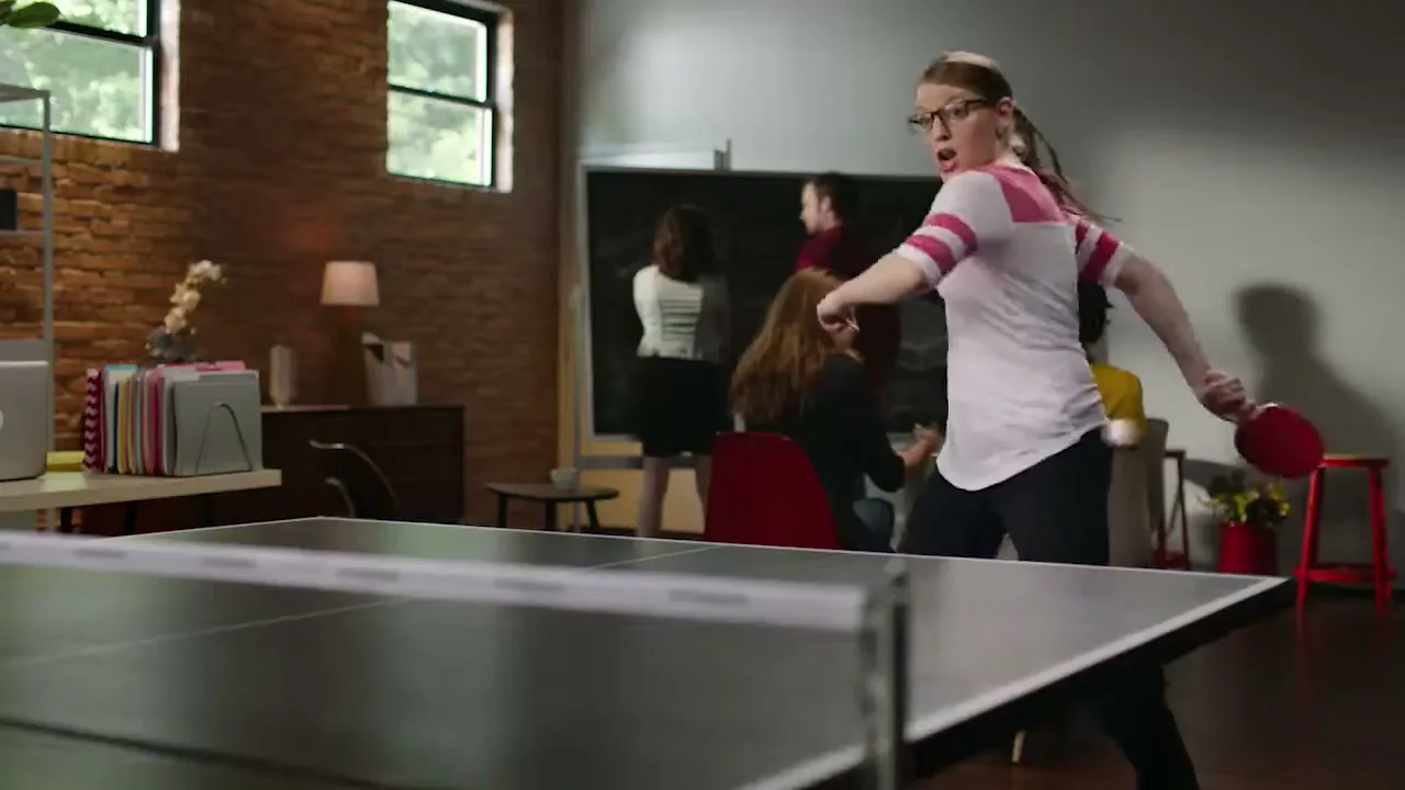 A young woman in glasses and a white and pink shirt plays ping pong in an office, with other colleagues in the background.